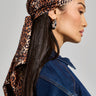 A person with long, dark hair is wearing the CHEETAH ANIMAL HEAD SCARF tied around their head. They are dressed in a sleeveless denim top and large hoop earrings, looking to the side against a plain background.