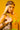 A person with long blonde hair and visible torso tattoos poses confidently against a bright yellow background, wearing a neon green bikini top, gold bangles, a CHEETAH ANIMAL HEAD SCARF hair accessory, and holding a matching purse.