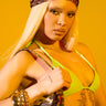 A person with long blonde hair and visible torso tattoos poses confidently against a bright yellow background, wearing a neon green bikini top, gold bangles, a CHEETAH ANIMAL HEAD SCARF hair accessory, and holding a matching purse.