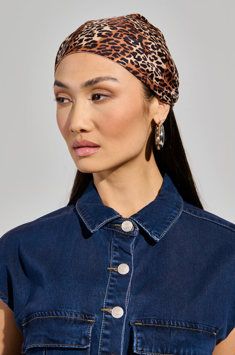 A woman wearing a chic CHEETAH ANIMAL HEAD SCARF and large hoop earrings looks to the side. She is dressed in a denim button-up shirt with short sleeves. The plain background highlights her calm expression and casual outfit.