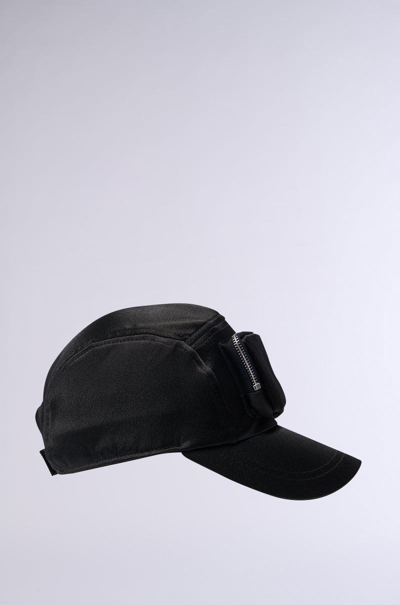 The SATIN LUXURY CARGO SNAPBACK is a black curved brim baseball cap with a subtle, sleek design and minimal detailing. It features a small zipper pocket on the side, blending seamlessly with the fabric for a modern, functional look. An adjustable Velcro strap ensures a perfect fit. The plain light gray background highlights the cap prominently. 3