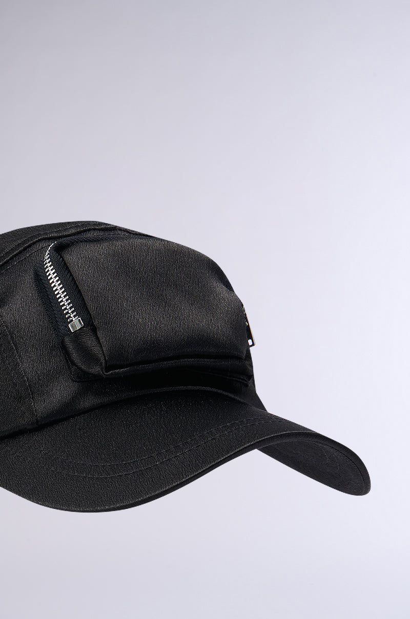 The SATIN LUXURY CARGO SNAPBACK is a black baseball cap with a textured surface, featuring a distinctive zipped pocket on the front. The metallic zipper contrasts strikingly with the cap's dark material. This curved brim snapback also includes an adjustable Velcro strap for a perfect fit. A plain light background highlights every detail of this elegant design. 4