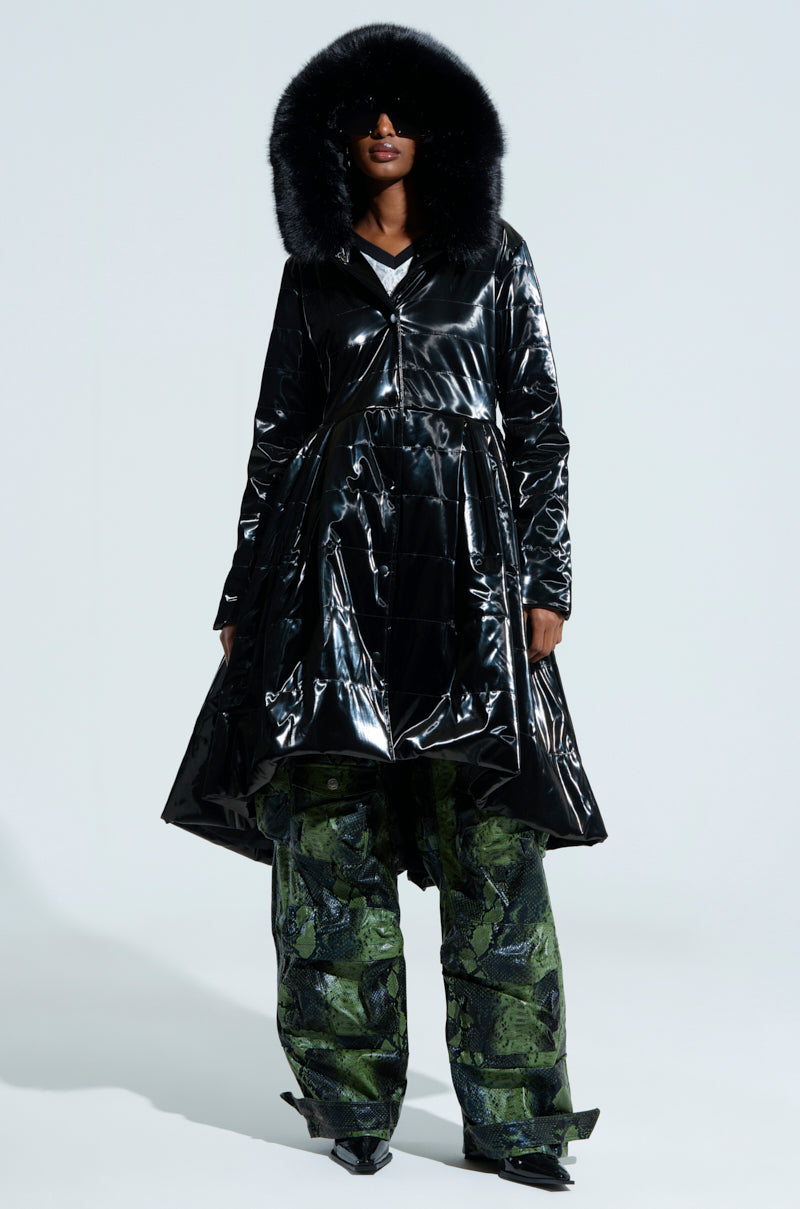 A person dons the MIDTOWN LIQUID VELVET PEPLUM COAT, teamed with oversized green patterned pants. They accessorize with black sunglasses, standing against a plain background.