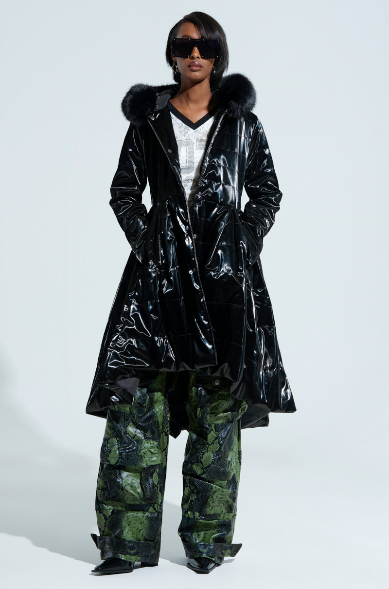 A person confidently stands against a plain background with hands in pockets, wearing the MIDTOWN LIQUID VELVET PEPLUM COAT featuring a shiny black finish and faux fur-trimmed hood, paired with sunglasses and green textured wide-leg pants, exuding a femme fatale allure.