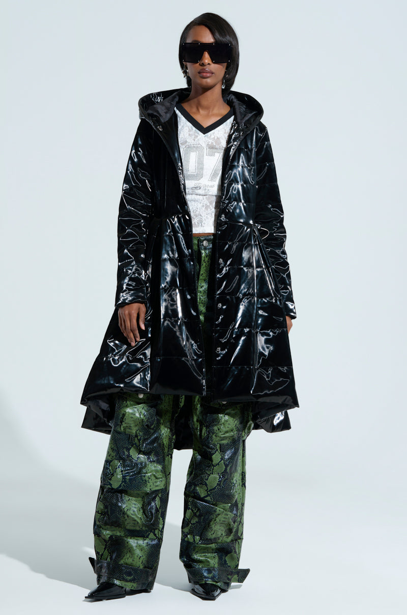 A person wearing the Midtown Liquid Velvet Peplum Coat over a white T-shirt with a large number, complemented by green snake-patterned wide-leg pants. They have short hair, dark sunglasses, and the scene is set against a plain light tone.