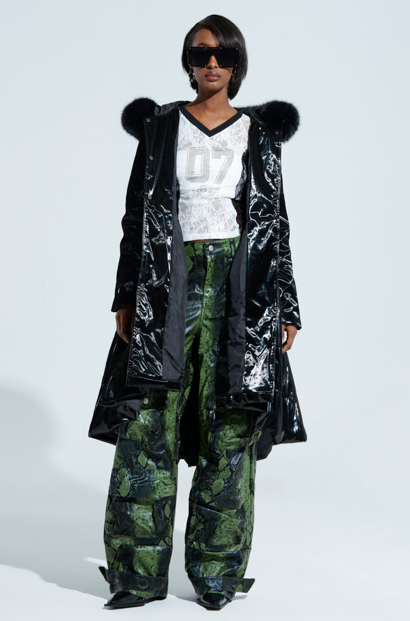 A person wearing the MIDTOWN LIQUID VELVET PEPLUM COAT with a faux fur trim and shiny finish, paired with a white top and green camouflage-patterned wide-leg pants. They have short hair and large black sunglasses in front of a plain light gray background.