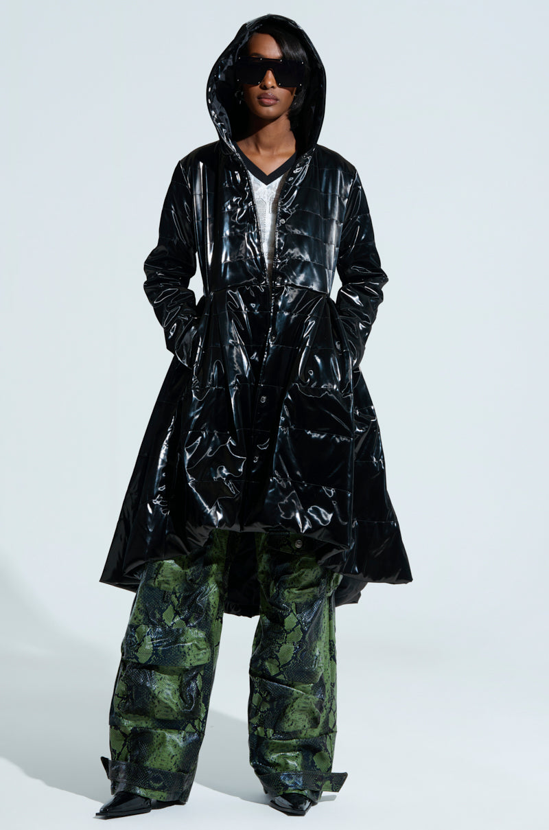 A person wearing a shiny black MIDTOWN LIQUID VELVET PEPLUM COAT with the hood up and green patterned pants stands confidently against a light background. They are sporting dark sunglasses and have their hands tucked into the coat pockets.