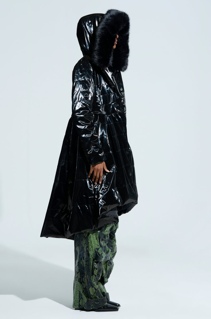 A person in the Midtown Liquid Velvet Peplum Coat, featuring a shiny black finish with faux fur trim, stands in profile. They are wearing green patterned pants and pointed shoes against a plain gray background.