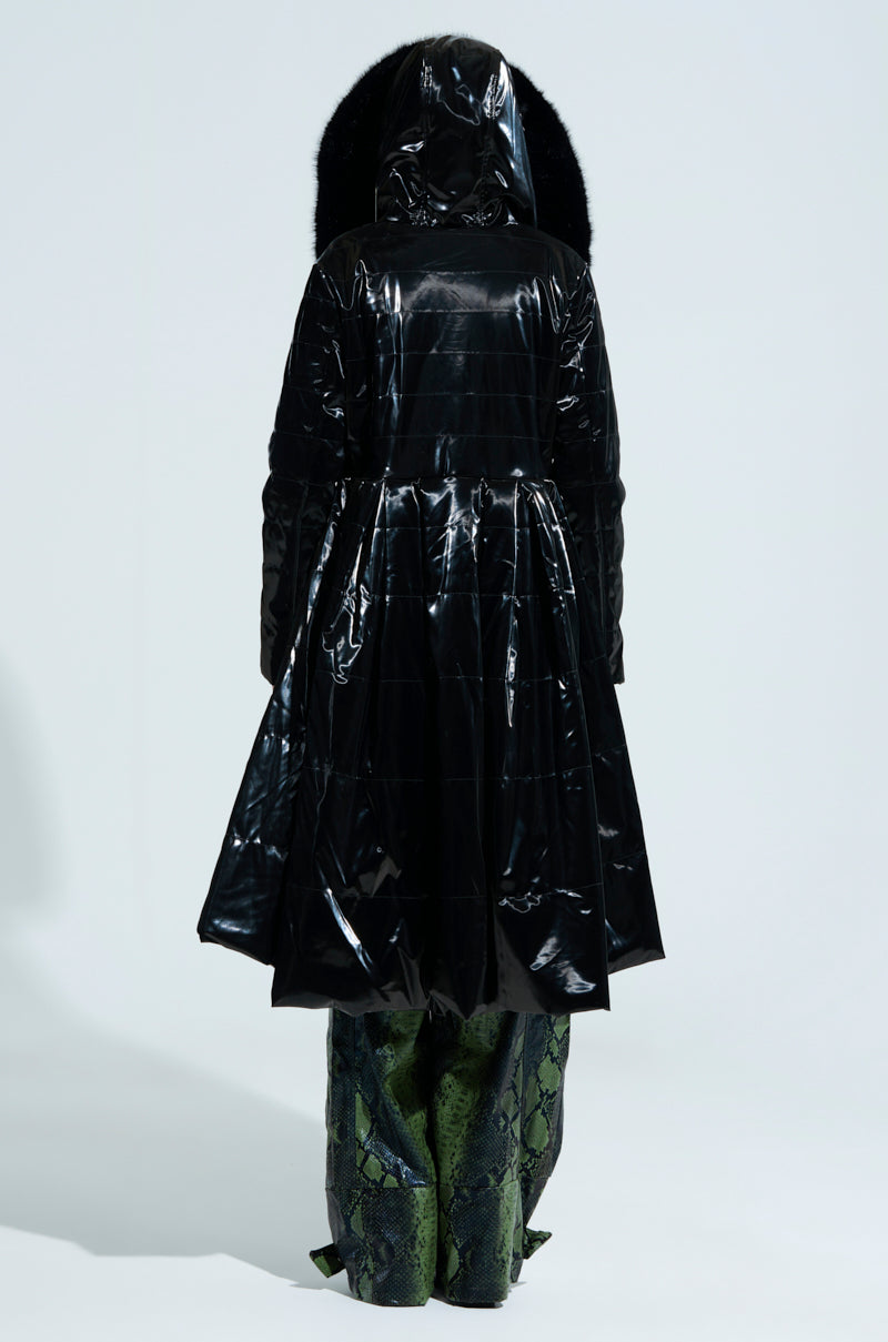 A person stands with their back to us, dressed in the dazzling MIDTOWN LIQUID VELVET PEPLUM COAT that features a flared bottom. The shiny coat makes a bold statement alongside their green snake-patterned pants against a simple white backdrop.