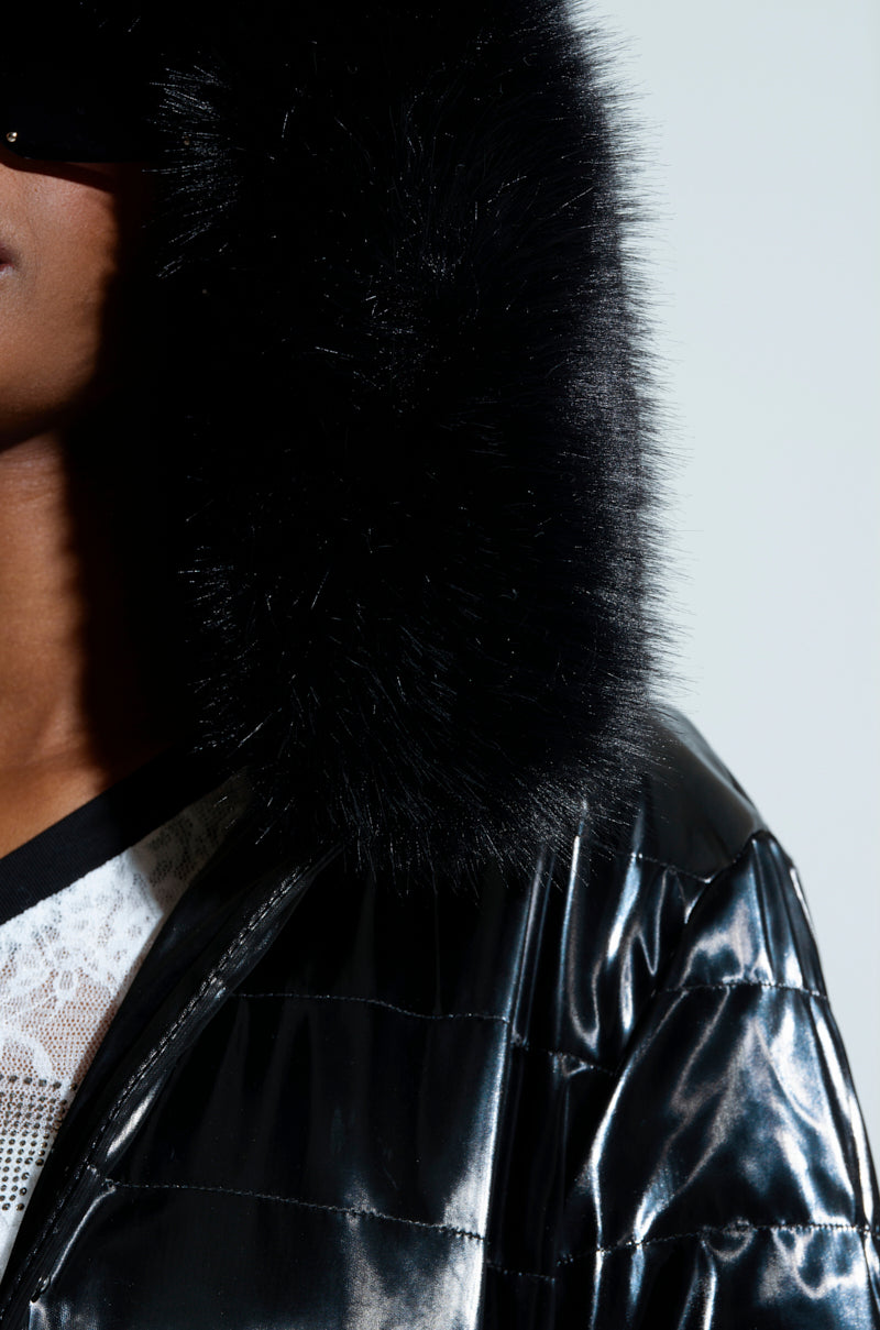 A close-up image showcases a person wearing a glossy black puffer jacket adorned with faux fur trim and a large, fluffy hood. The coat evokes the style of the Midtown Liquid Velvet Peplum Coat and is paired with a lace top underneath. The simple background highlights the texture of the fabric.