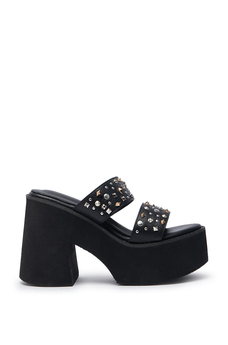 The AZALEA WANG MIKAEL BLACK CHUNKY SLIP ON SANDAL features black faux leather, two wide straps with gold & silver studs plus rhinestones, a platform sole, chunky high heel, and open toe design.
