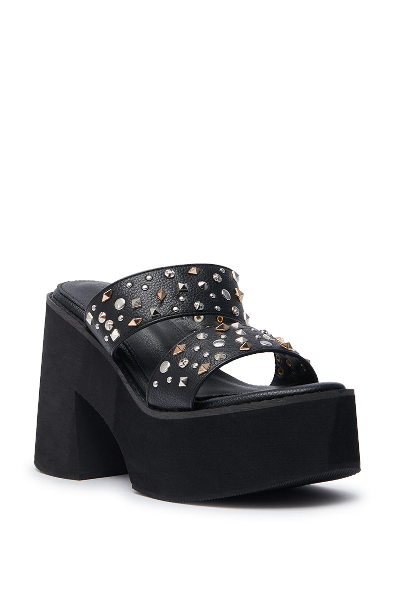 The AZALEA WANG MIKAEL BLACK CHUNKY SLIP ON SANDAL features a black faux leather platform design with a high chunky heel and two wide straps decorated with gold and silver metal studs in assorted shapes.