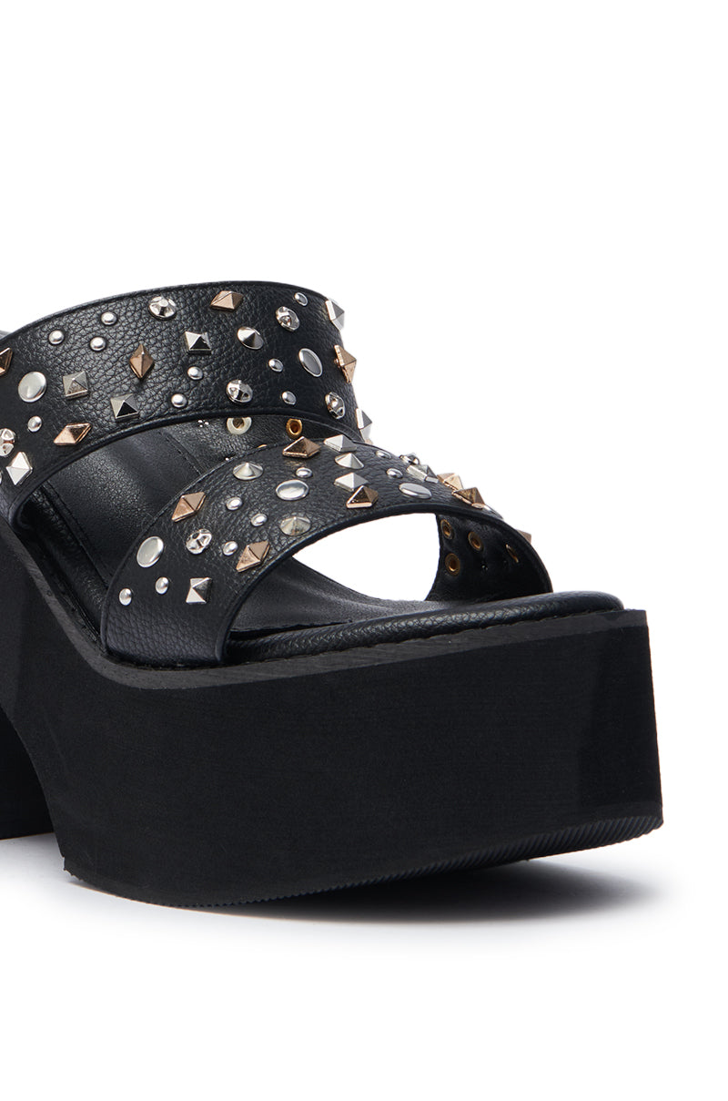The AZALEA WANG MIKAEL BLACK CHUNKY SLIP ON SANDAL features a platform sole, two wide faux leather straps with silver and gold studs and rhinestones, shown close-up from the front and side against a white background.