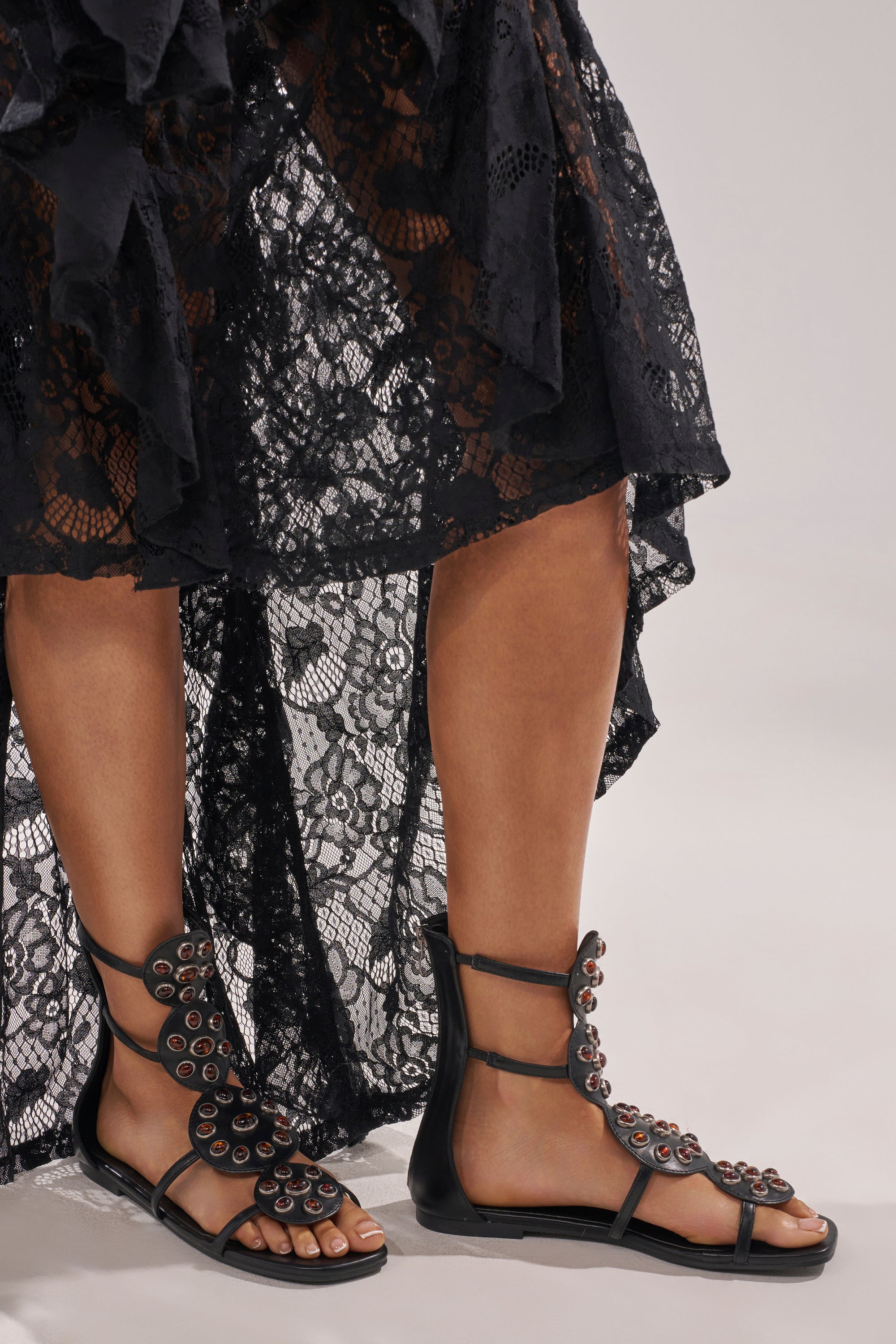 A person wearing a black lace skirt and AZALEA WANG MIKAERE Black Brown Stone Embellished Flat Strap Sandals stands on a light gray surface. 1
