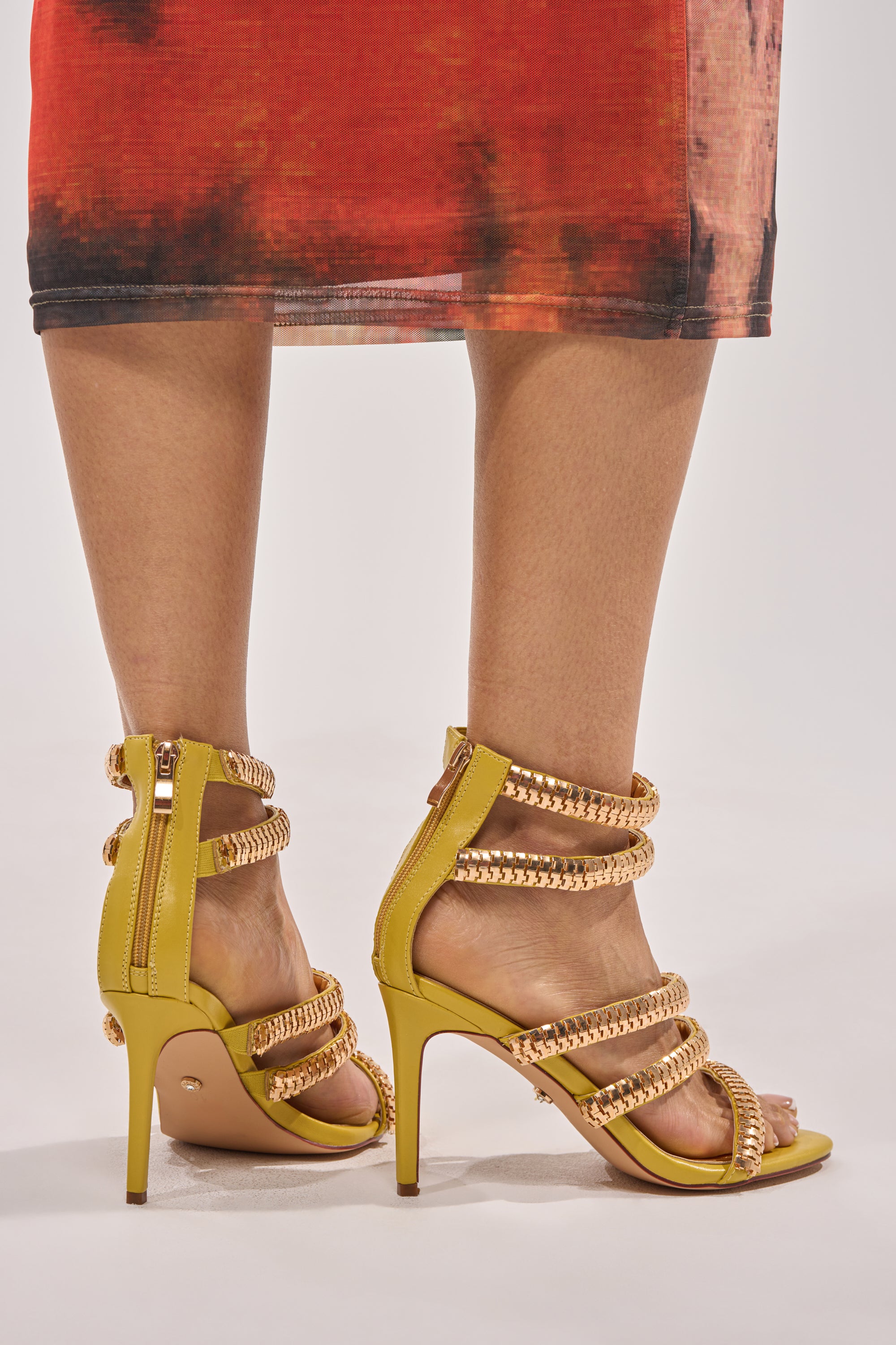 A person wears the AZALEA WANG MILA CITRON YELLOW GOLD HARDWARE EMBELLISHED STRAPPY STILETTO SANDAL with a red and black tie-dye skirt. The photo highlights the standout party shoes' gold chain straps from the back and side at ankle level. 2