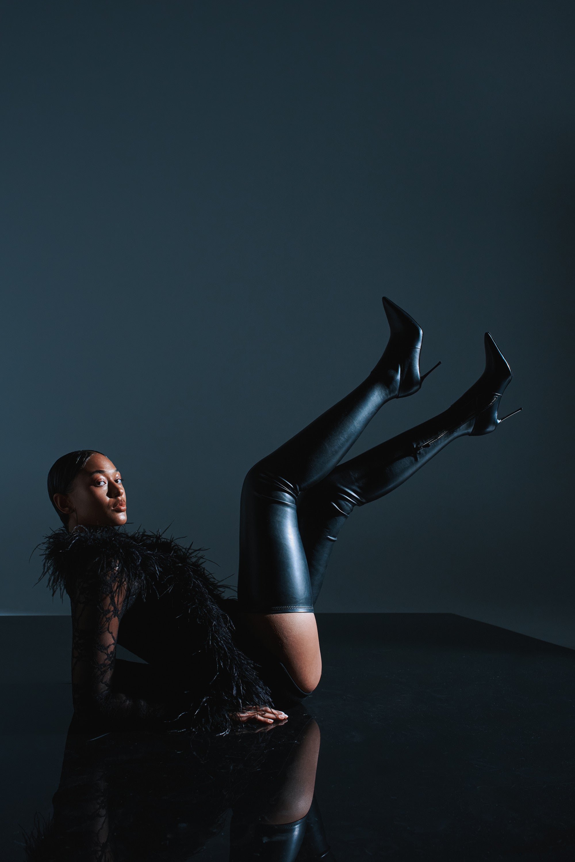 A woman poses confidently on her back against a dark background, wearing a black feathered top, faux leather shorts, and the AZALEA WANG MILANDRA BLACK THIGH HIGH SEXY BOOT, her legs raised and bent at the knees.
