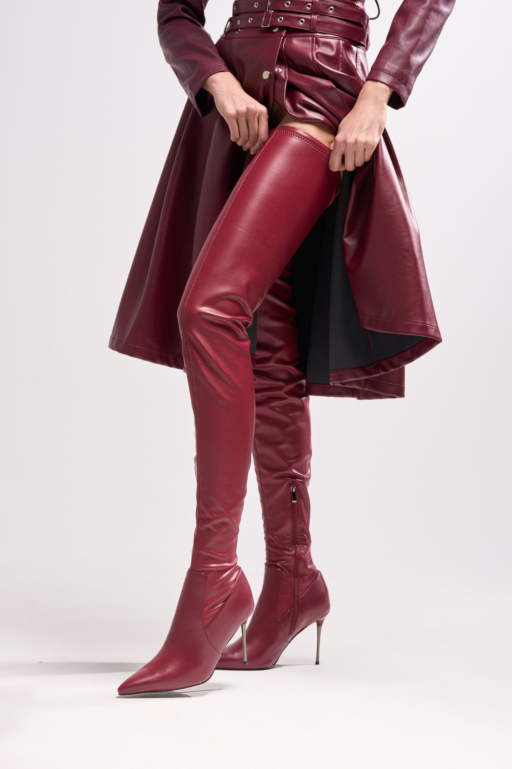 Wearing the AZALEA WANG MILANDRA BURGUNDY STRETCH PU THIGH HIGH BOOT and a burgundy leather coat with metallic accents, a person stands against a plain background, adjusting the shiny boot.