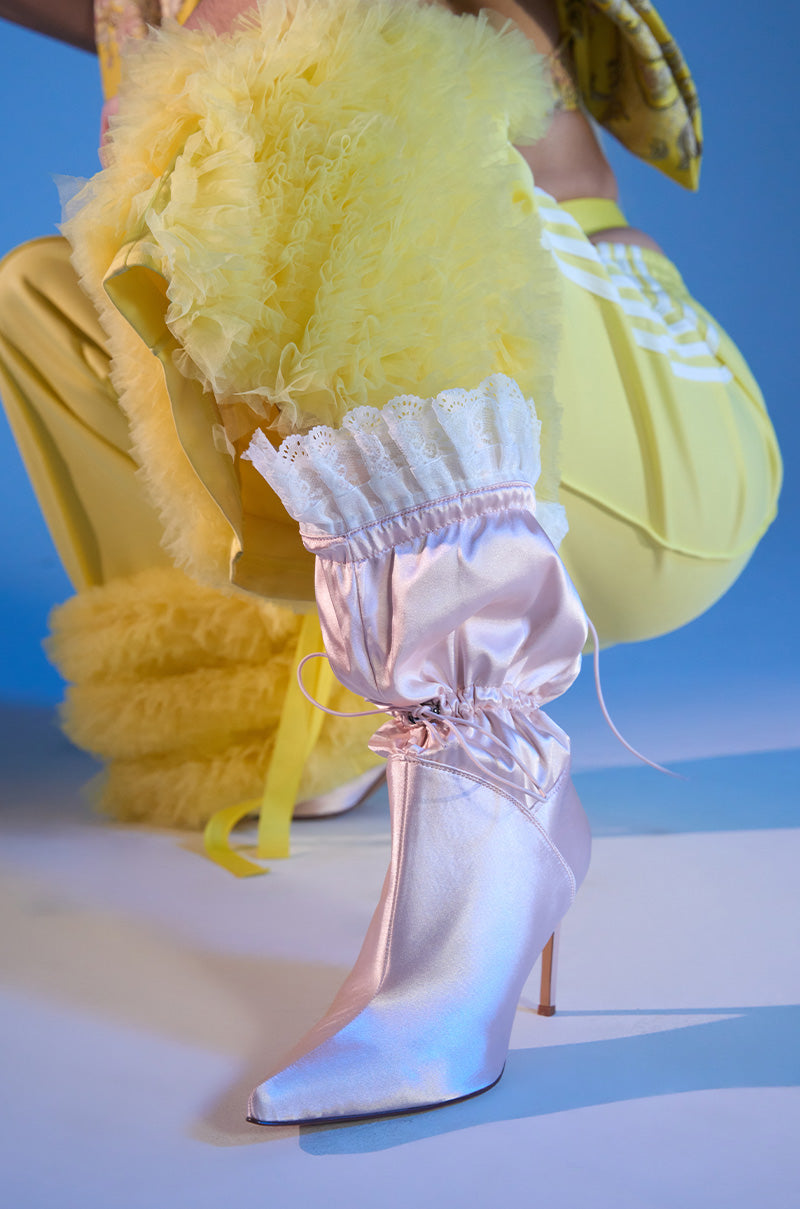 Squatting against a blue backdrop, a person in bright yellow ruffled pants and top showcases the AZALEA WANG MILDRED PINK CINCHING BOOTIE with white lace trim, gathered details, and a chic pointed toe.