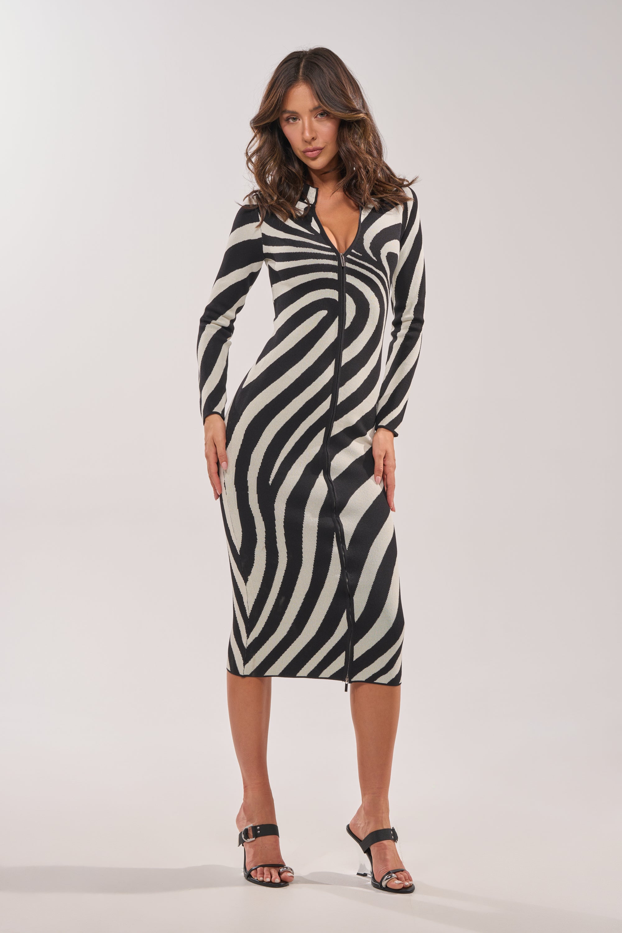 A woman models the UNDER MY SPELL MIDI DRESS, a black and white striped knit bodycon style with a front zipper, paired with black open-toe heels, posing confidently against a simple light background. 8