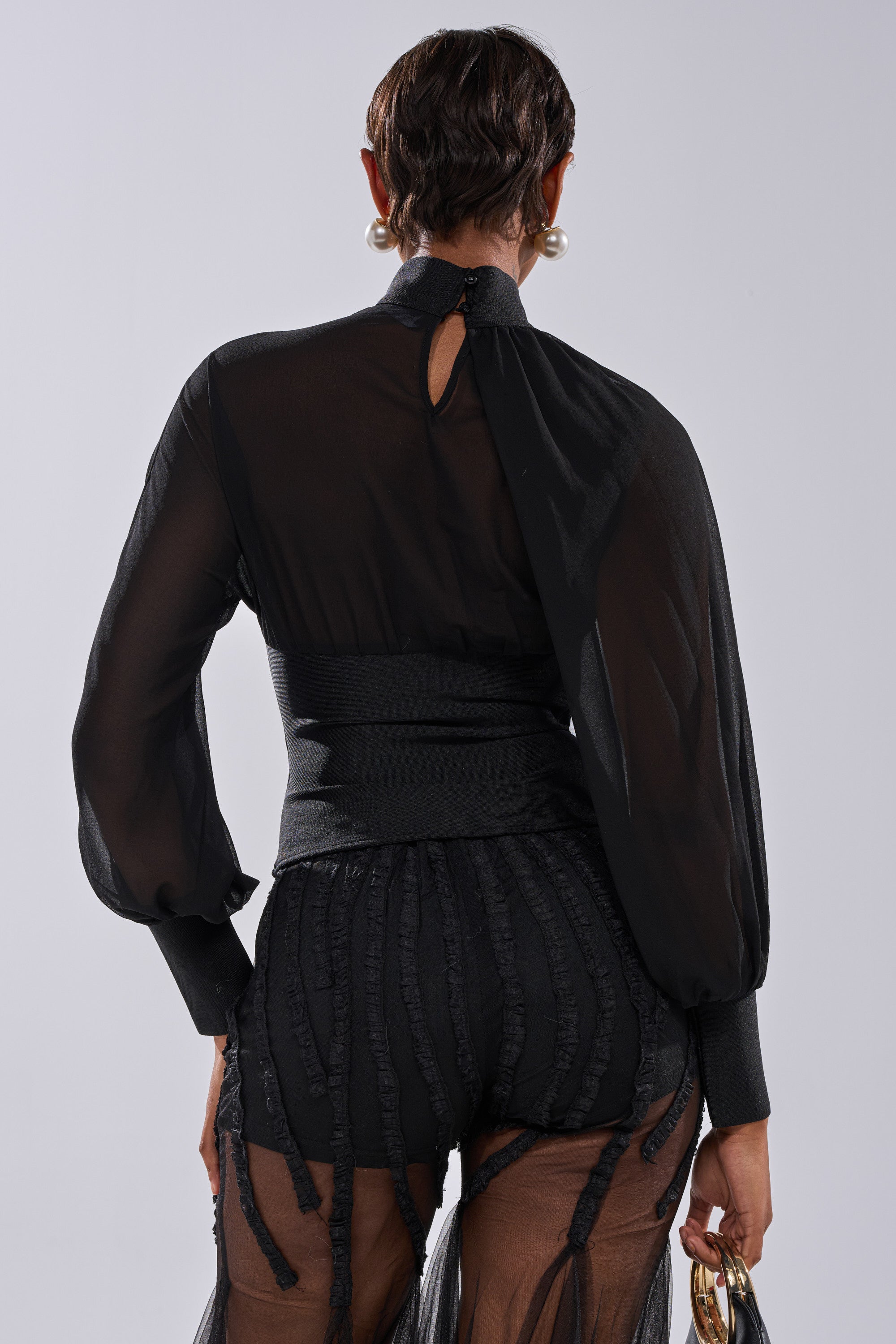 A person with short hair and large pearl earrings is shown from the back wearing the FIRST THING FIRST BLOUSE—a sheer black chiffon blouse—paired with matching high-waisted sheer pants against a light gray background.