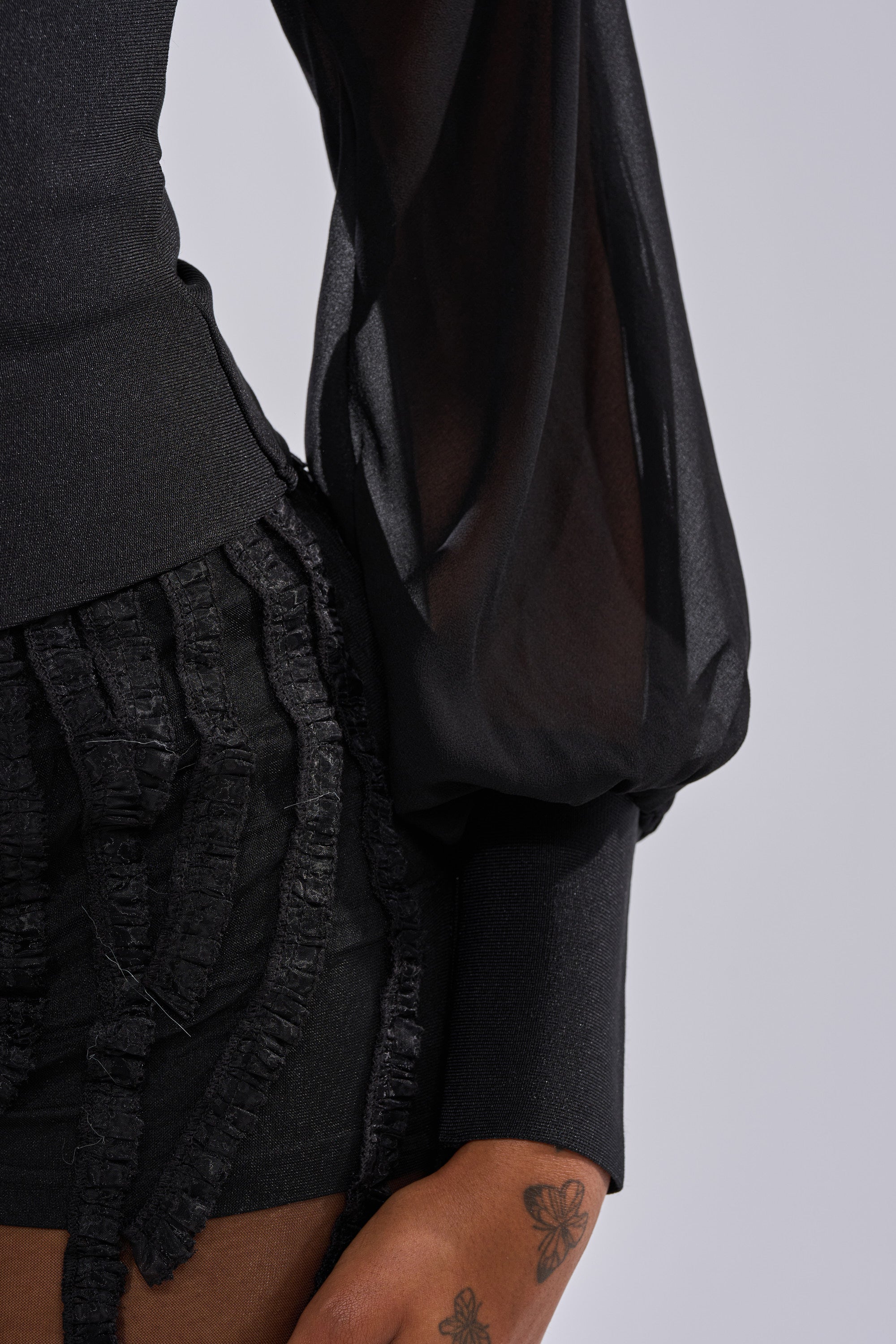 A close-up of a person wearing the FIRST THING FIRST BLOUSE, a sheer black chiffon top, with textured black shorts. Their hand, featuring a butterfly tattoo, rests by their side against a plain light background.