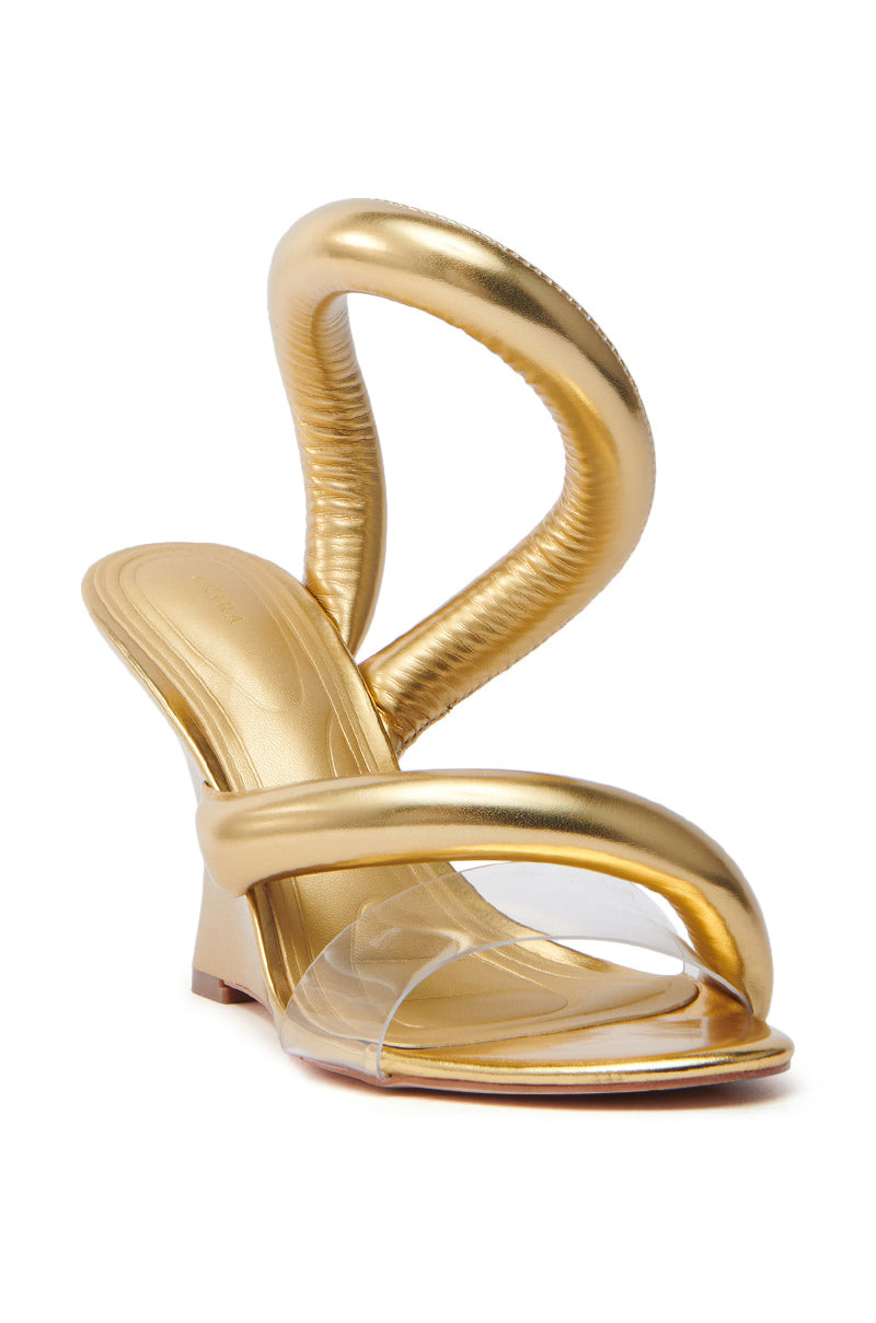 The AKIRA MINORU GOLD WEDGE SANDAL features a gold high-heel with an artistic, twisting strap design and clear front strap. Made from metallic faux leather, it has an open almond toe and sculpted heel, offering a modern, avant-garde look.
