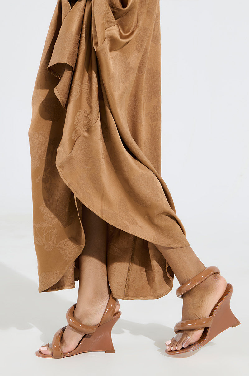 A person in a brown patterned skirt is walking, displaying only their legs and feet. They're wearing AKIRA MINORU NUDE WEDGE SANDALS with a comfort cushion insole. The soft gray background adds a minimalistic and elegant touch.