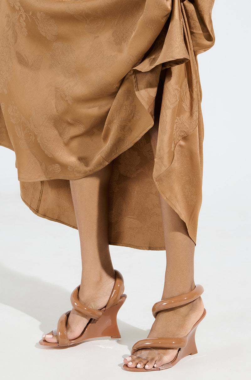 The image shows a person wearing a brown, textured knee-length skirt and the AKIRA Minoru nude wedge sandals with clear straps and a comfort cushion insole, standing on a light-colored floor.