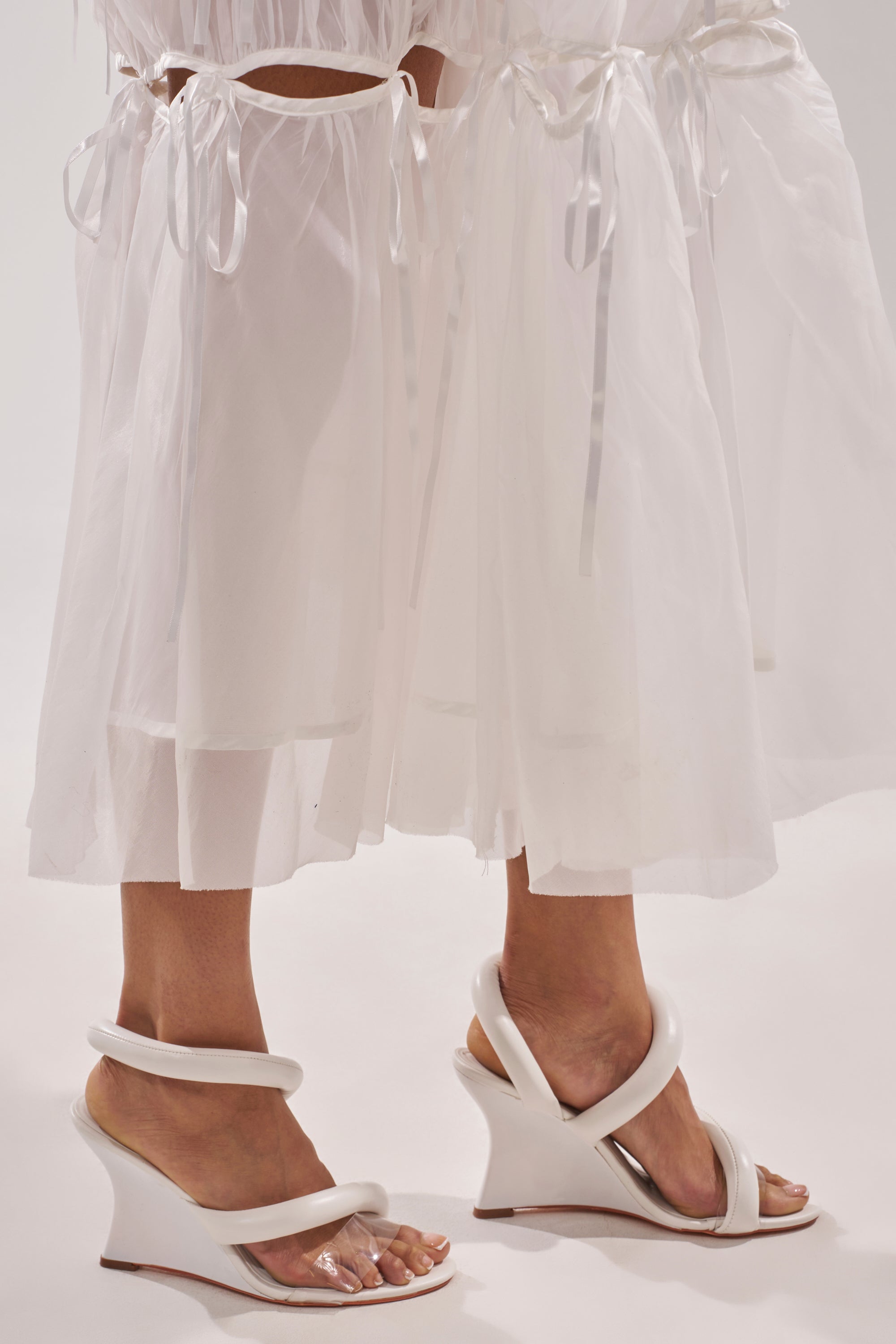 Two people stand side by side in sheer white skirts with ribbon ties, both wearing the AKIRA MINORU WHITE WEDGE SANDAL, which features curved modern straps, a transparent front band, and clear accents, against a light background. 1
