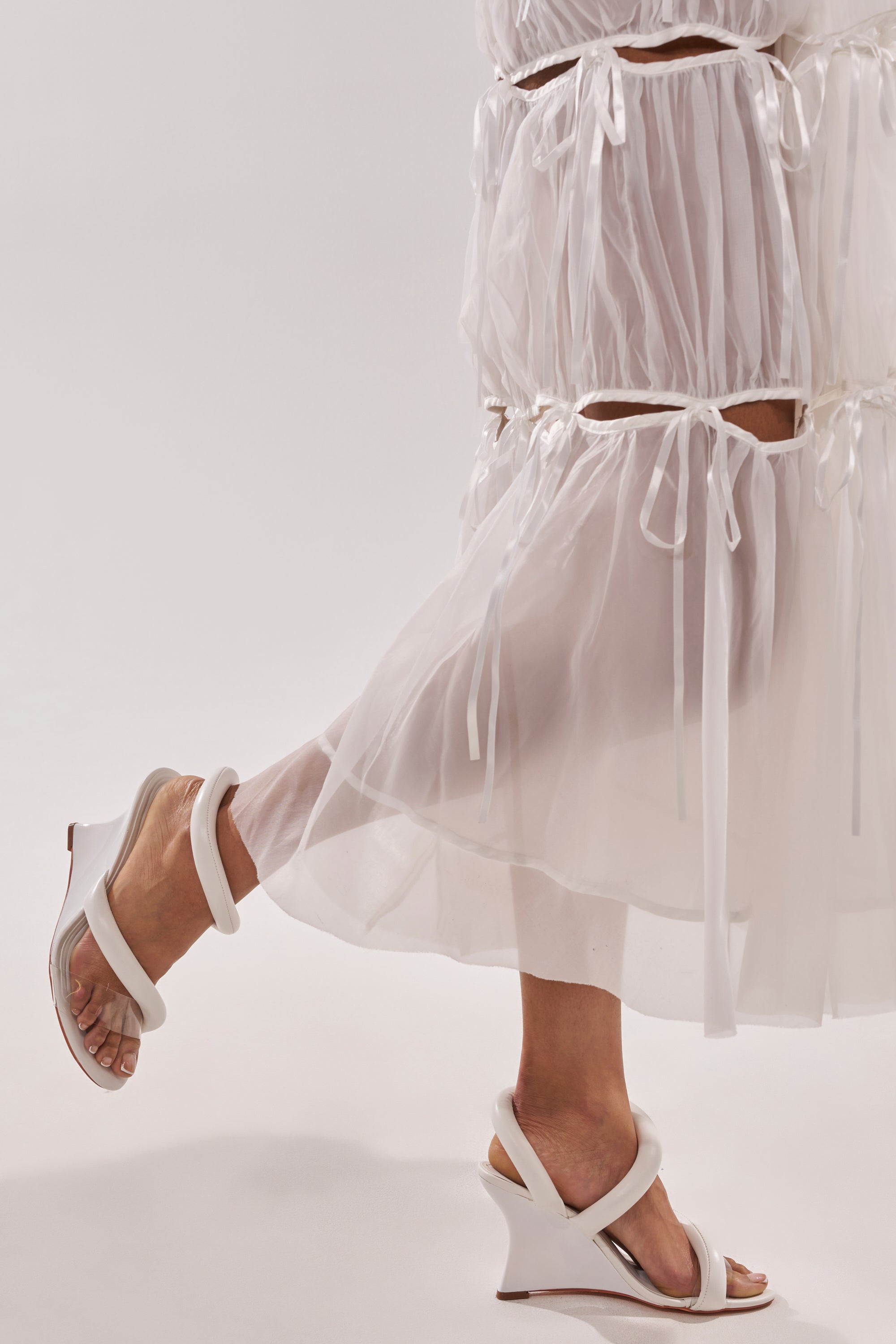 A person poses in a sheer white dress with ribbons, wearing AKIRA MINORU WHITE WEDGE SANDAL featuring a comfort cushion insole, shown from mid-calf down with one leg bent and the other foot flat against a light background. 2