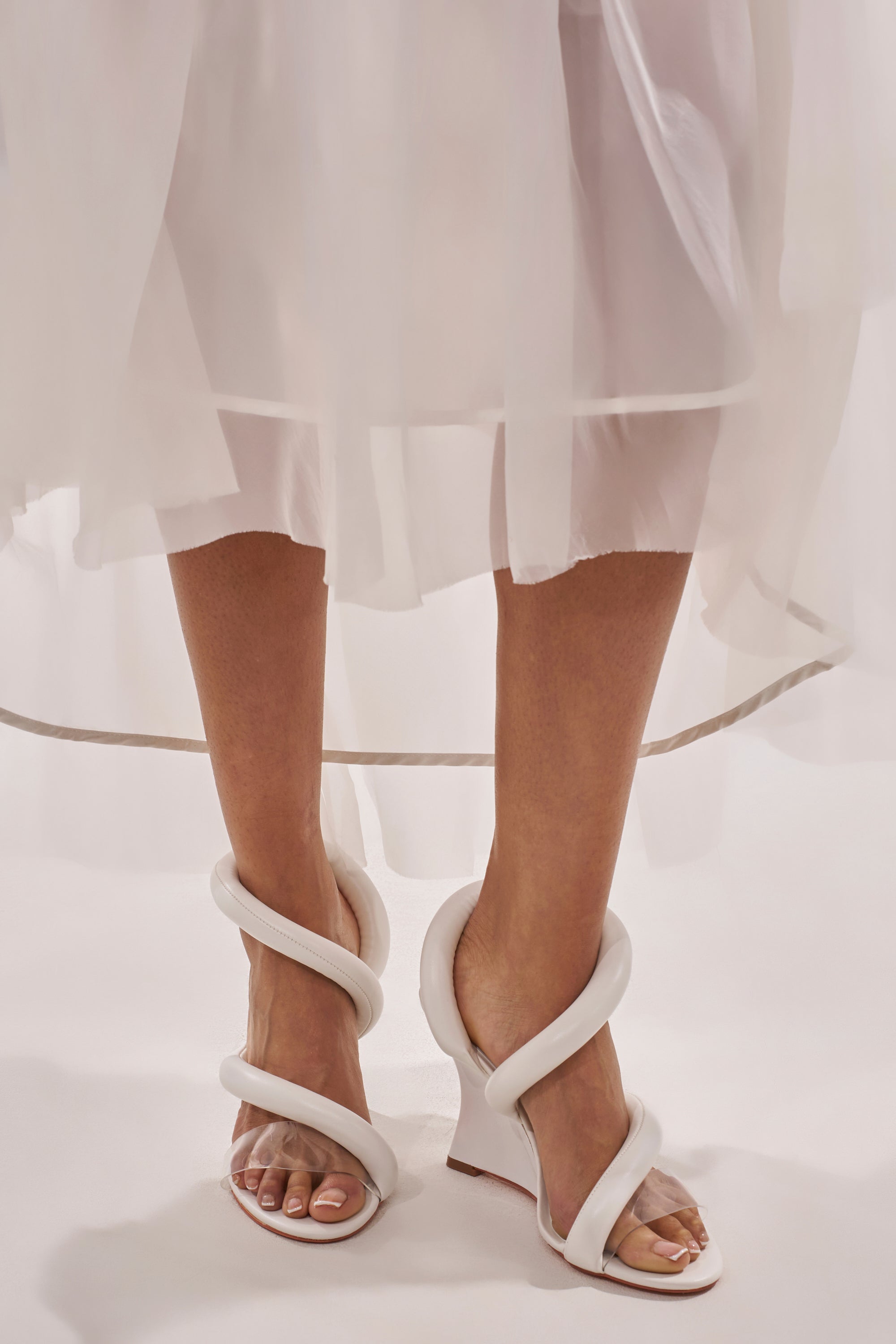 Wearing a sheer, white dress, a person stands on a light surface and showcases AKIRA MINORU WHITE WEDGE SANDALS, featuring thick sculptural straps spiraling around the ankle and a comfort cushion insole for all-day wear. 3