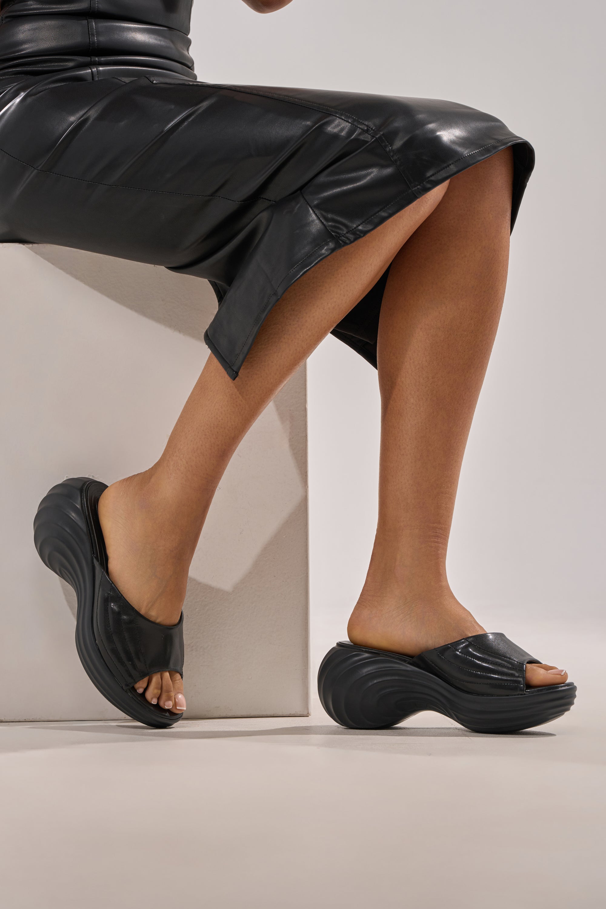 A person in a black leather skirt sits on a white block, highlighting their feet in AZALEA WANG MIRAGE BLACK PU FLATFORM SLIP ON SANDALS with wavy soles and comfort cushion insoles against a neutral background for a minimal look.