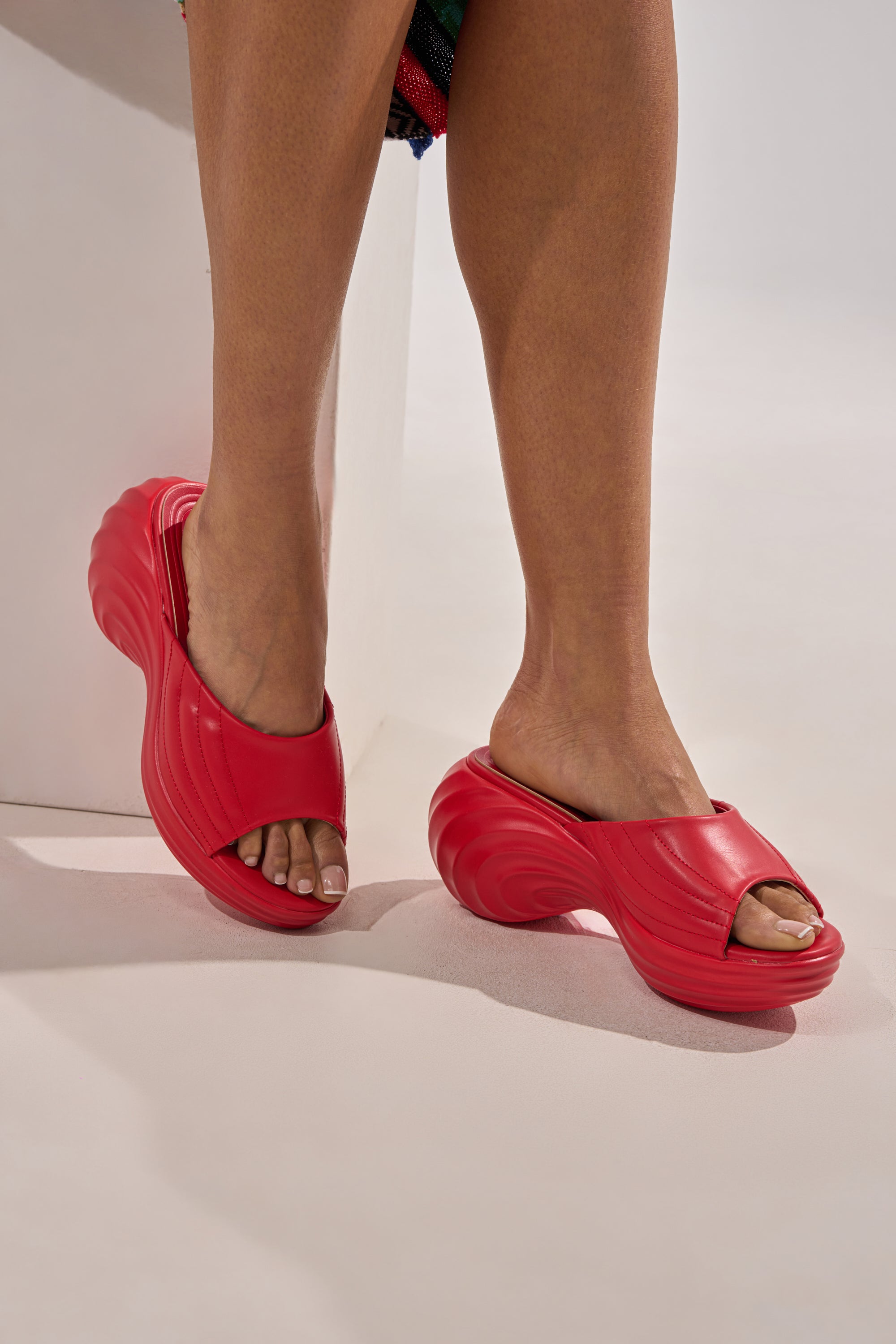 Someone showcases the bold, sculpted design of the AZALEA WANG MIRAGE RED PU FLATFORM SLIP ON SANDAL with its wave-inspired sole while standing on a light-colored surface.