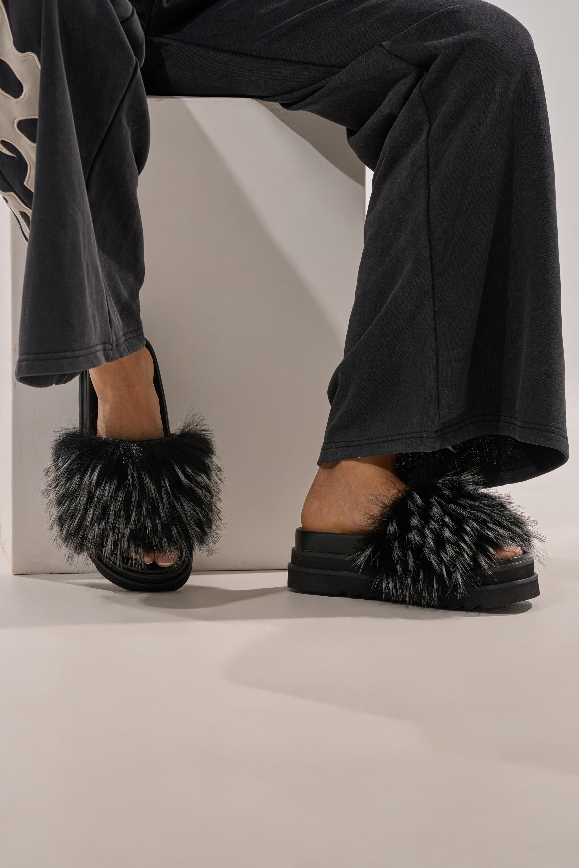 A person wearing black pants sits on a white block, showing their feet in the AKIRA MIRAY BLACK MULTI BLACK FUR SLIDE sandals with chunky platform soles and thick, fluffy black-and-gray faux fur straps. 0