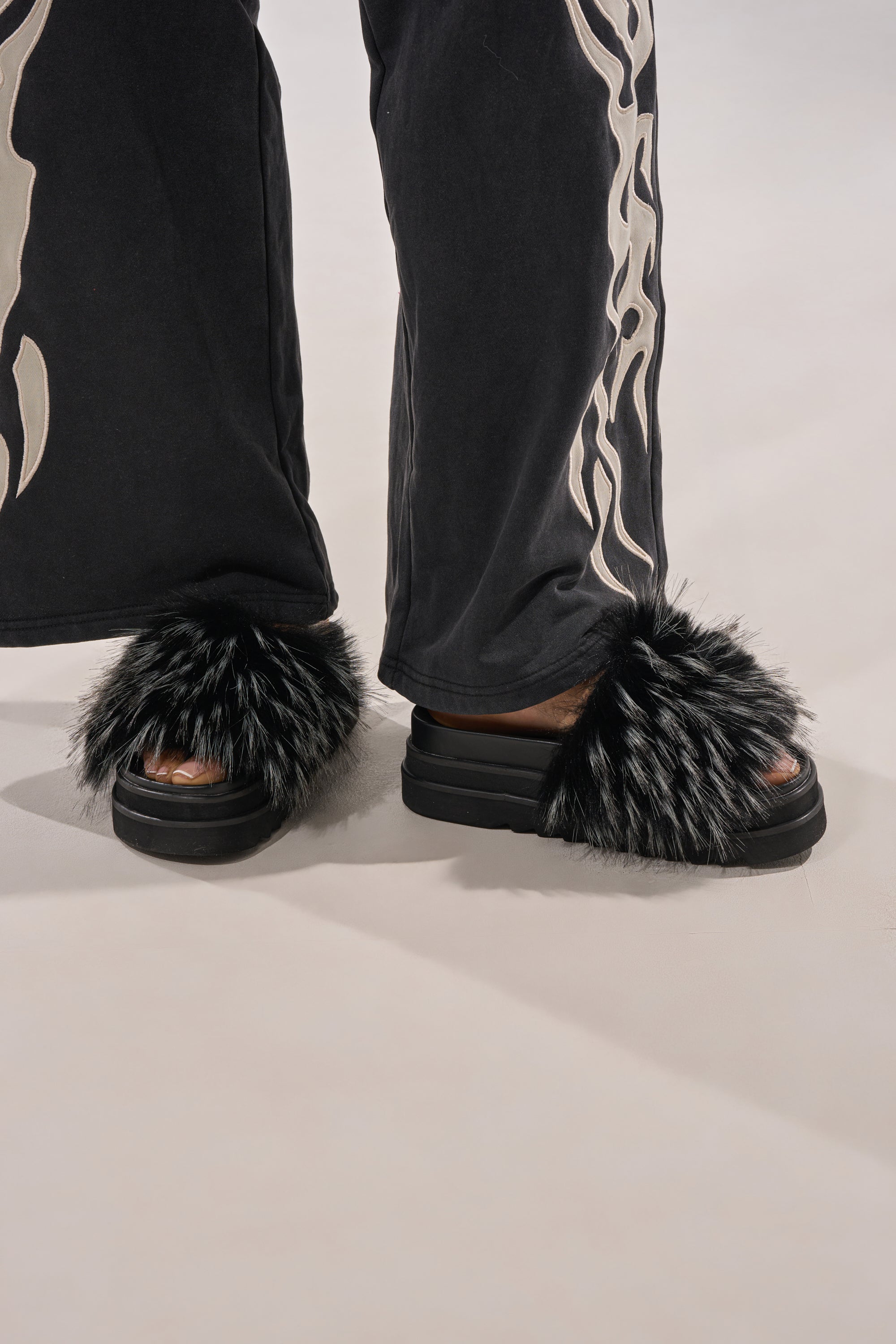 A person wearing black pants with white abstract patterns and the AKIRA MIRAY BLACK MULTI BLACK FUR SLIDE sandals with a chunky platform sole, standing on a light-colored surface. 1