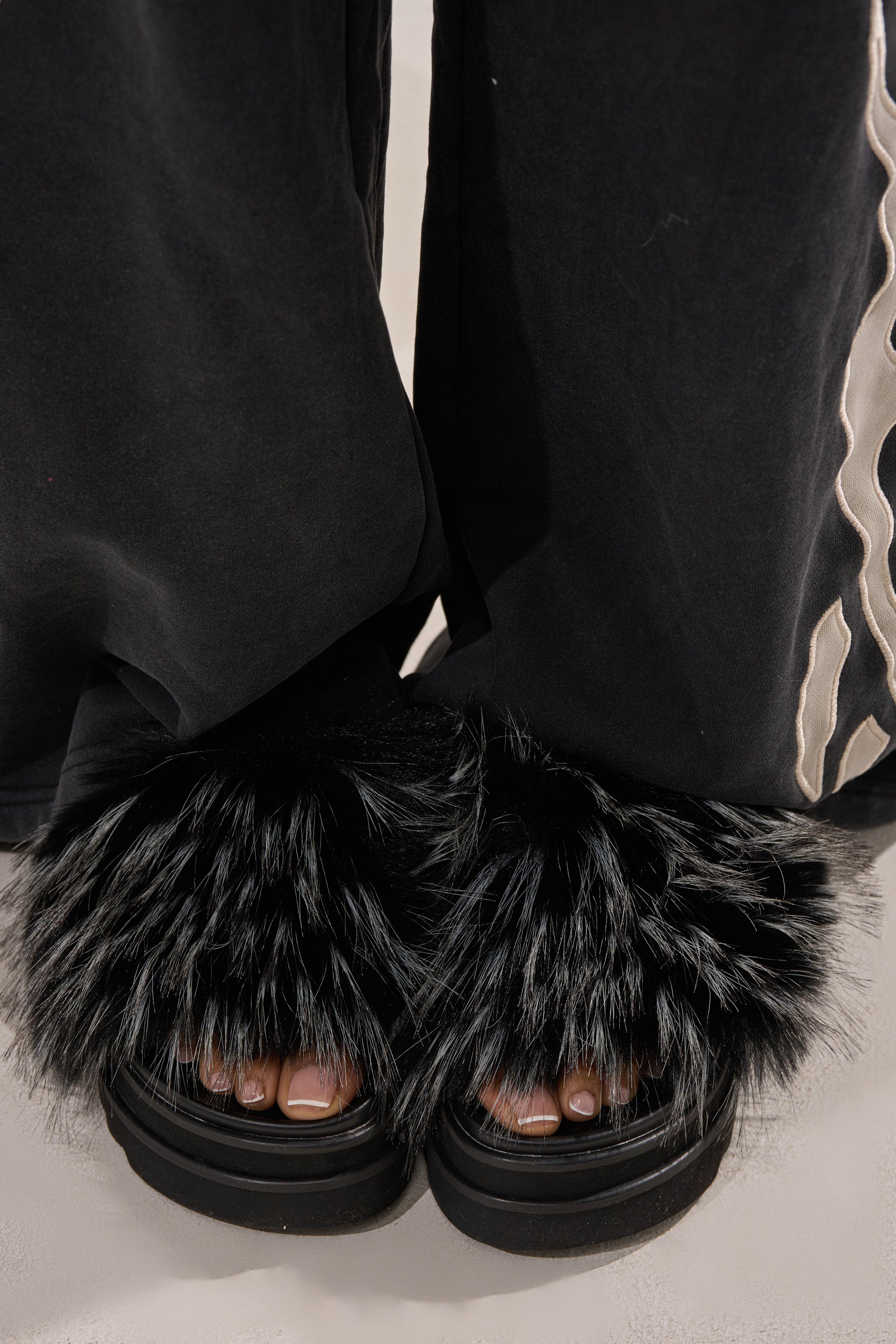 A person wears black pants with a white wavy stripe and flaunts their toes in the AKIRA MIRAY BLACK MULTI BLACK FUR SLIDES, featuring fluffy black faux fur and a chunky platform sole. 2