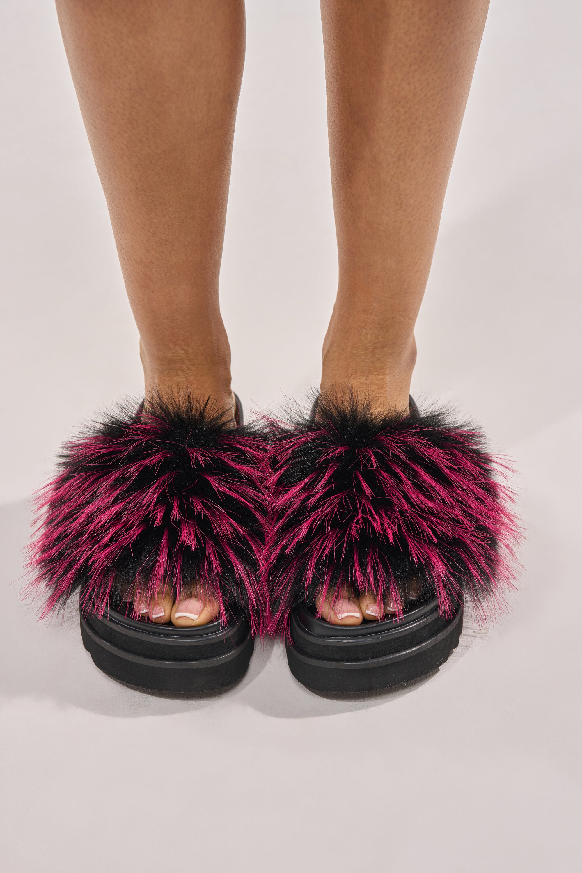 A person wears the AKIRA MIRAY PINK MULTI FUR SLIDE—black faux leather platform sandals with spiky pink faux fur in a bold Y2K slide style; only their lower legs and feet show against a plain background. 1