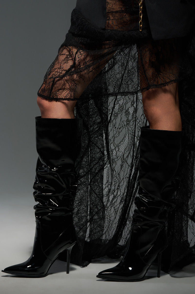 In a studio setting, an individual stands poised, wearing the AZALEA WANG MITICA BLACK PATENT SLOUCH BOOT paired with a sheer black lace skirt. The sleek faux patent leather knee-high boots feature stiletto heels, and the dark background enhances the outfit's textures and elegance, creating an alluring presence.