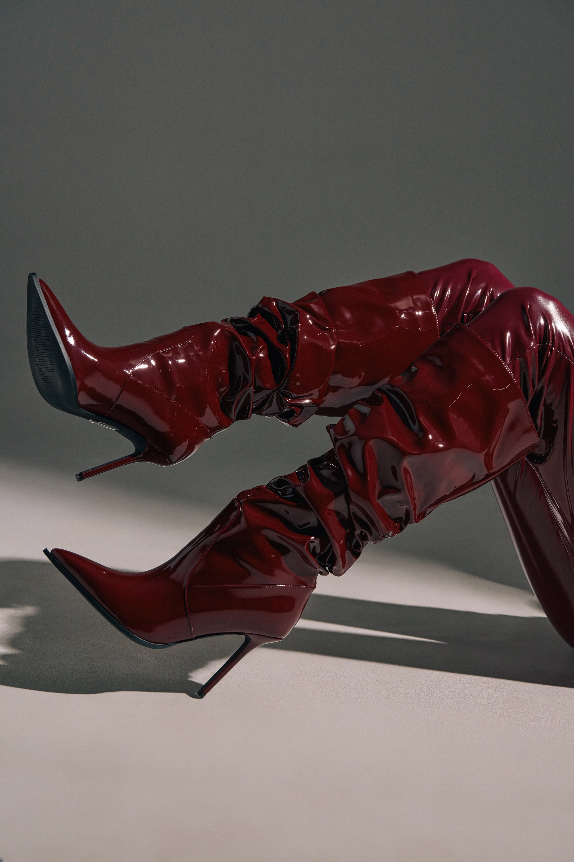 The AZALEA WANG MITICA BURGUNDY PATENT SLOUCH BOOT features shiny dark red faux patent leather, ruched detailing, and stiletto heels, shown on a model posing with crossed legs against a neutral background.