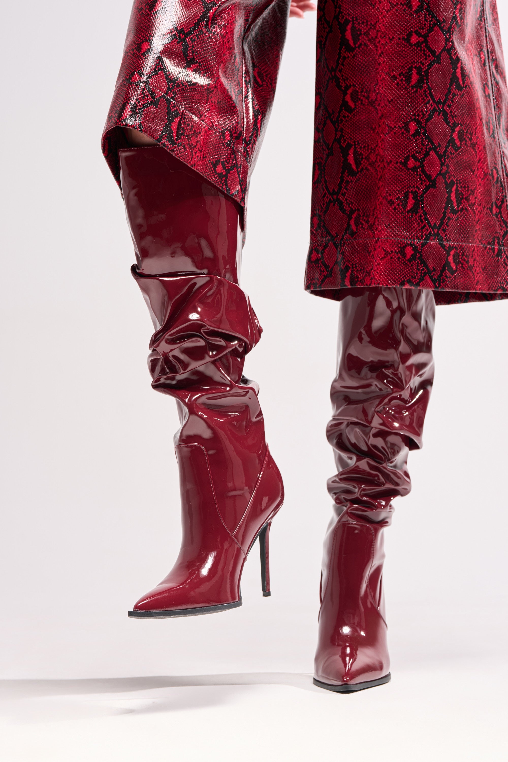 A person wearing the AZALEA WANG MITICA BURGUNDY PATENT SLOUCH BOOT with knee-high shine and stiletto heels, paired with a red snakeskin-patterned skirt or coat, stands against a light background.