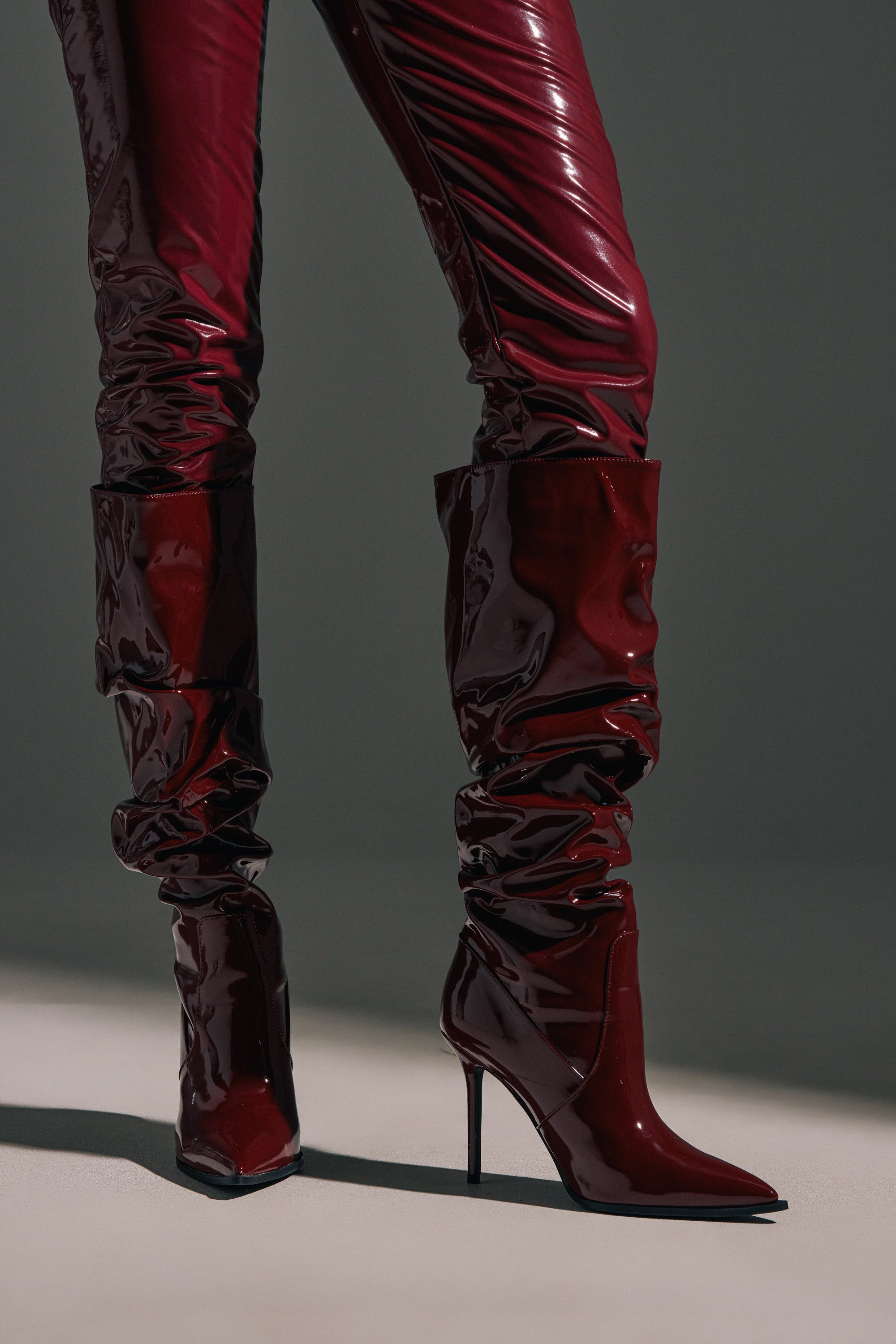 A person wearing AZALEA WANG MITICA BURGUNDY PATENT SLOUCH BOOTS and glossy faux patent leather pants stands on a light surface, casting a shadow behind them.