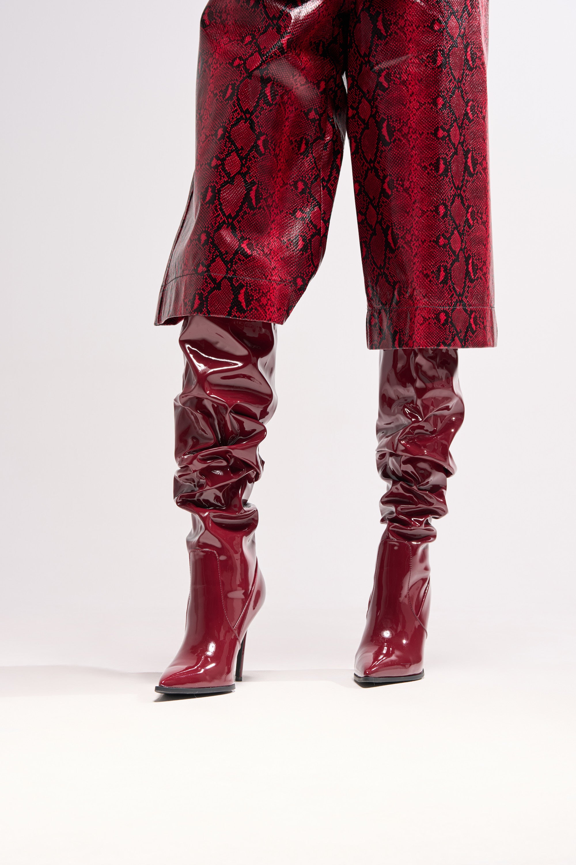 A person wearing the AZALEA WANG MITICA BURGUNDY PATENT SLOUCH BOOT with stiletto heels and red faux snakeskin pants, shown from mid-thigh down against a plain light background.