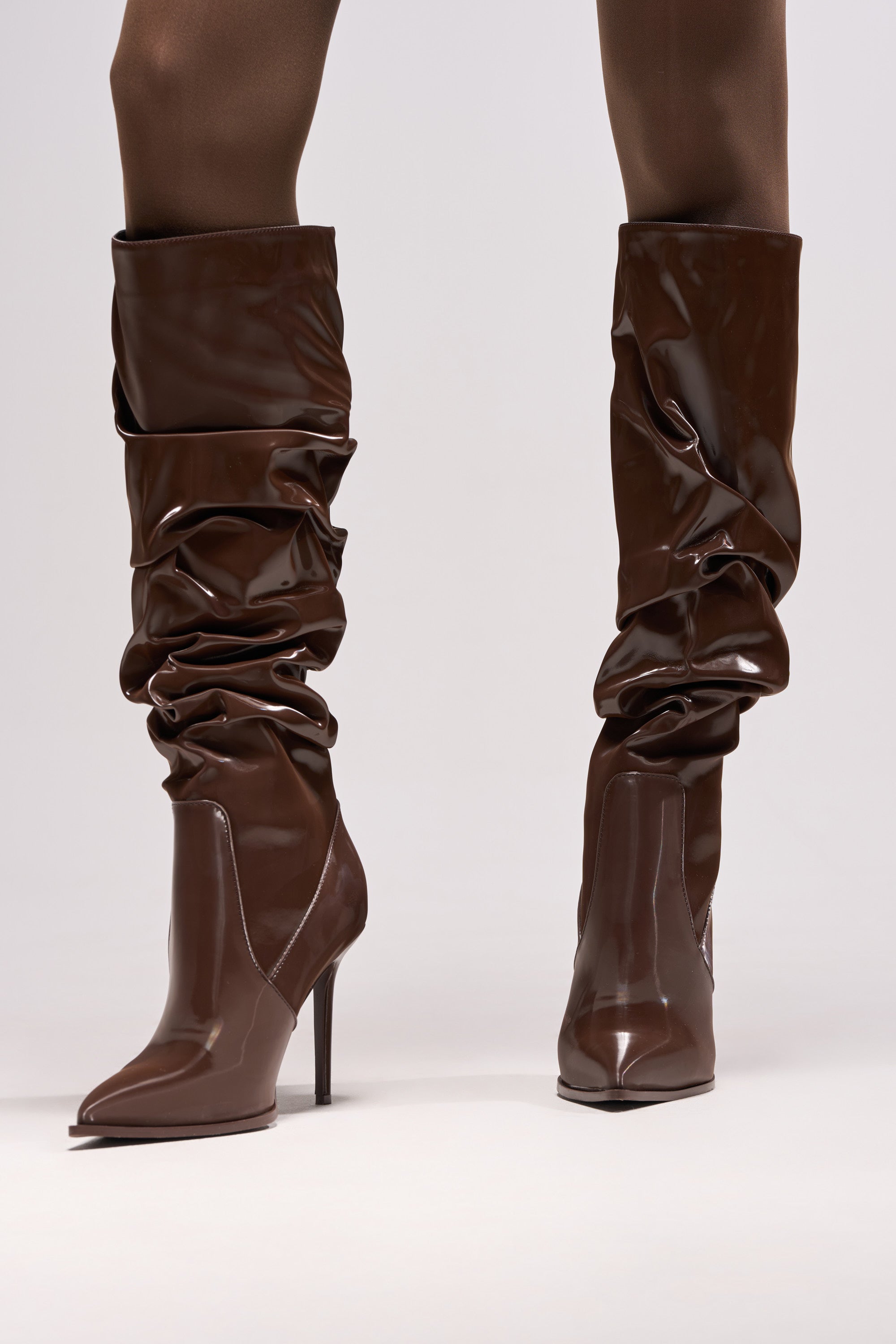 Close-up of a person wearing the AZALEA WANG MITICA DARK BROWN PATENT RUCHED SEXY BOOT—knee-high, stiletto heel, and shiny finish—styled with dark tights against a plain background.