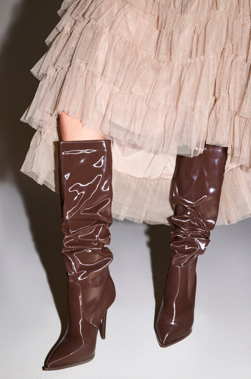 A person in a ruffled beige tulle skirt and AZALEA WANG MITICA DARK BROWN PATENT RUCHED SEXY BOOT with a slouchy, knee-high silhouette stands on a light gray surface.
