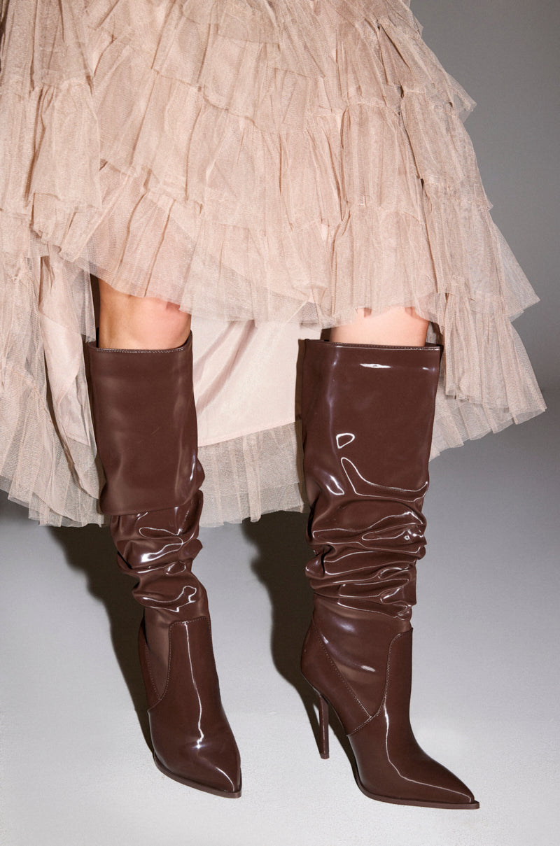 A person stands on a gray surface wearing a light beige ruffled tulle skirt and the AZALEA WANG MITICA DARK BROWN PATENT RUCHED SEXY BOOT—a shiny, knee-high, stiletto-heeled boot.