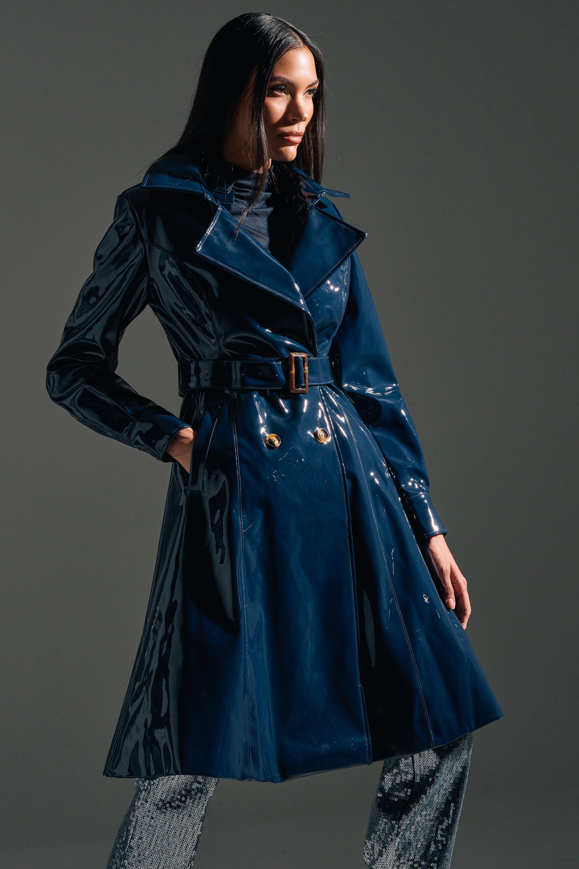 A woman poses confidently against a gray background in the MITICA PATENT TRENCH IN NAVY from AKIRA Label, styled over a black top and textured pants.