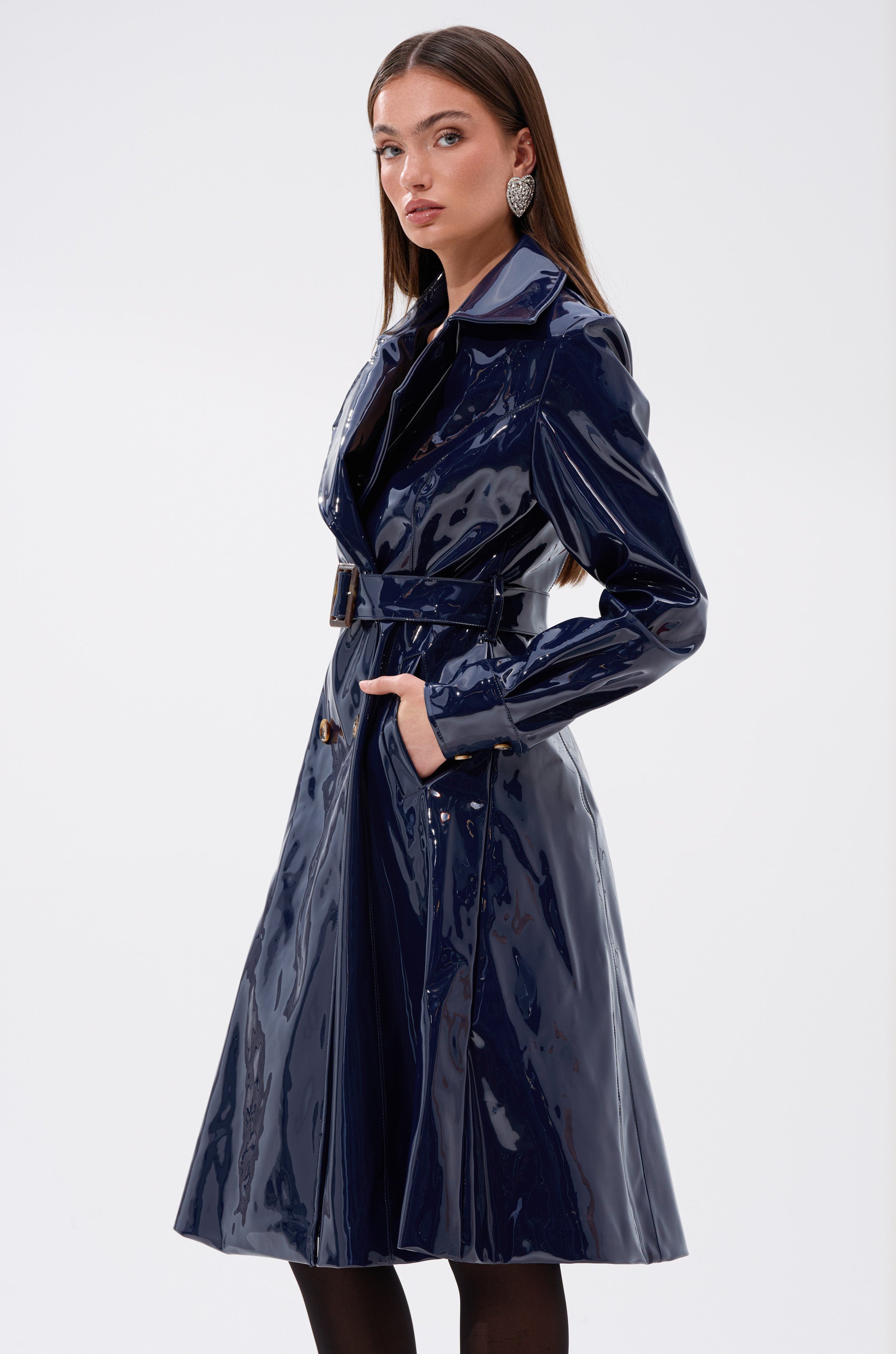 A woman models the MITICA PATENT TRENCH IN NAVY—a shiny faux leather trench with a belted waist—paired with black tights and statement earrings, posing against a plain white background with one hand in her pocket.