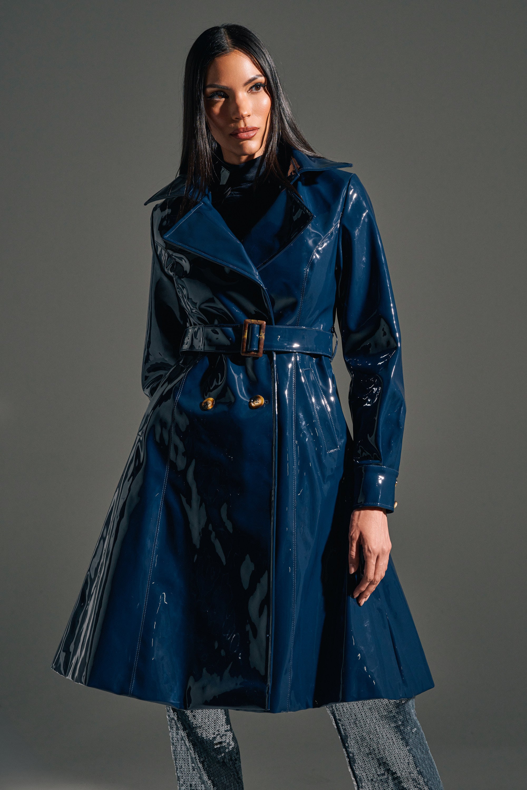 A woman poses confidently against a plain gray background, wearing the MITICA PATENT TRENCH IN NAVY with a longline silhouette over a black top and gray pants.
