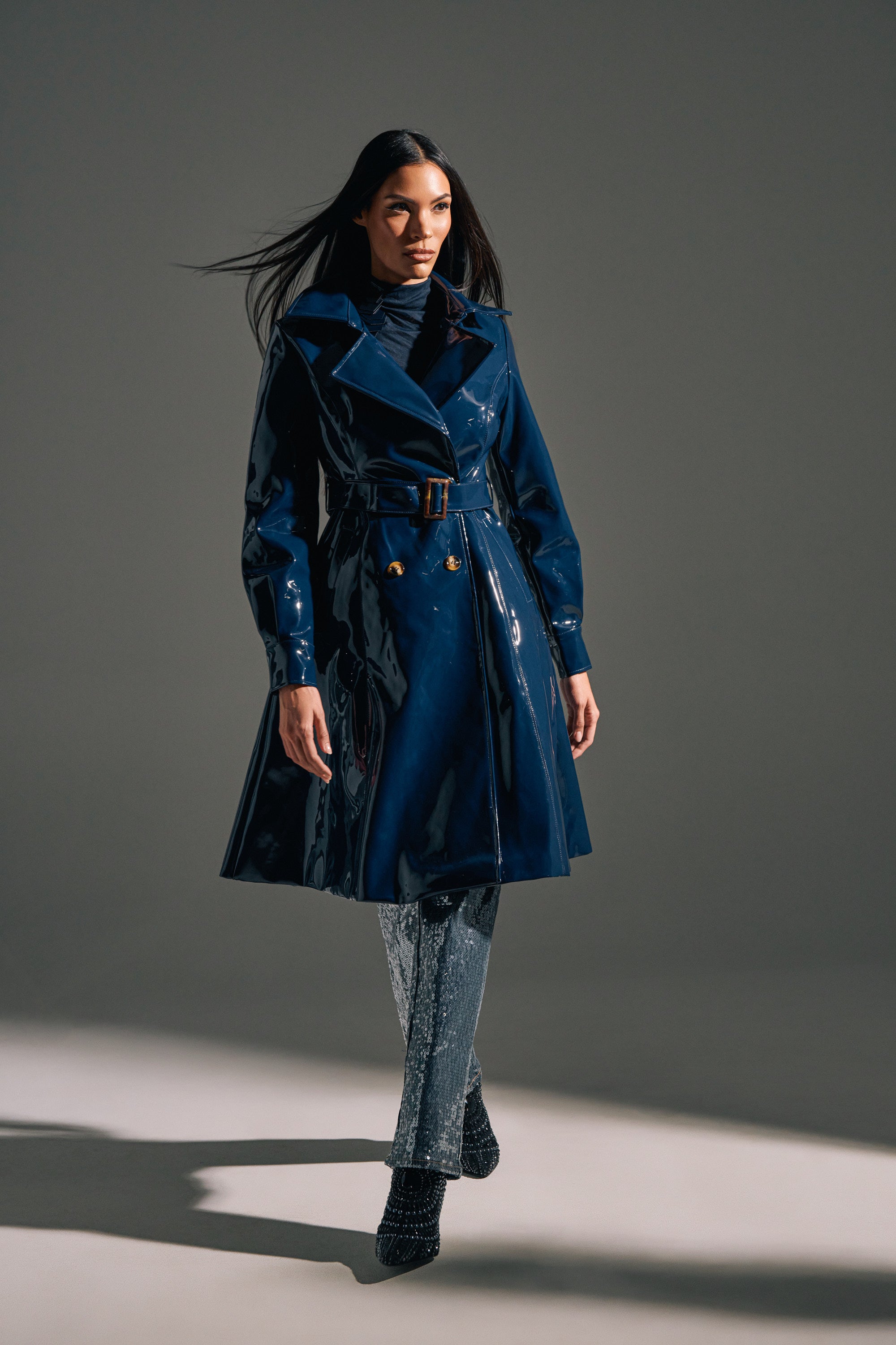 A woman walks confidently in the MITICA PATENT TRENCH IN NAVY, a glossy longline belted trench coat, styled with sparkly silver pants and black boots against a plain gray background.