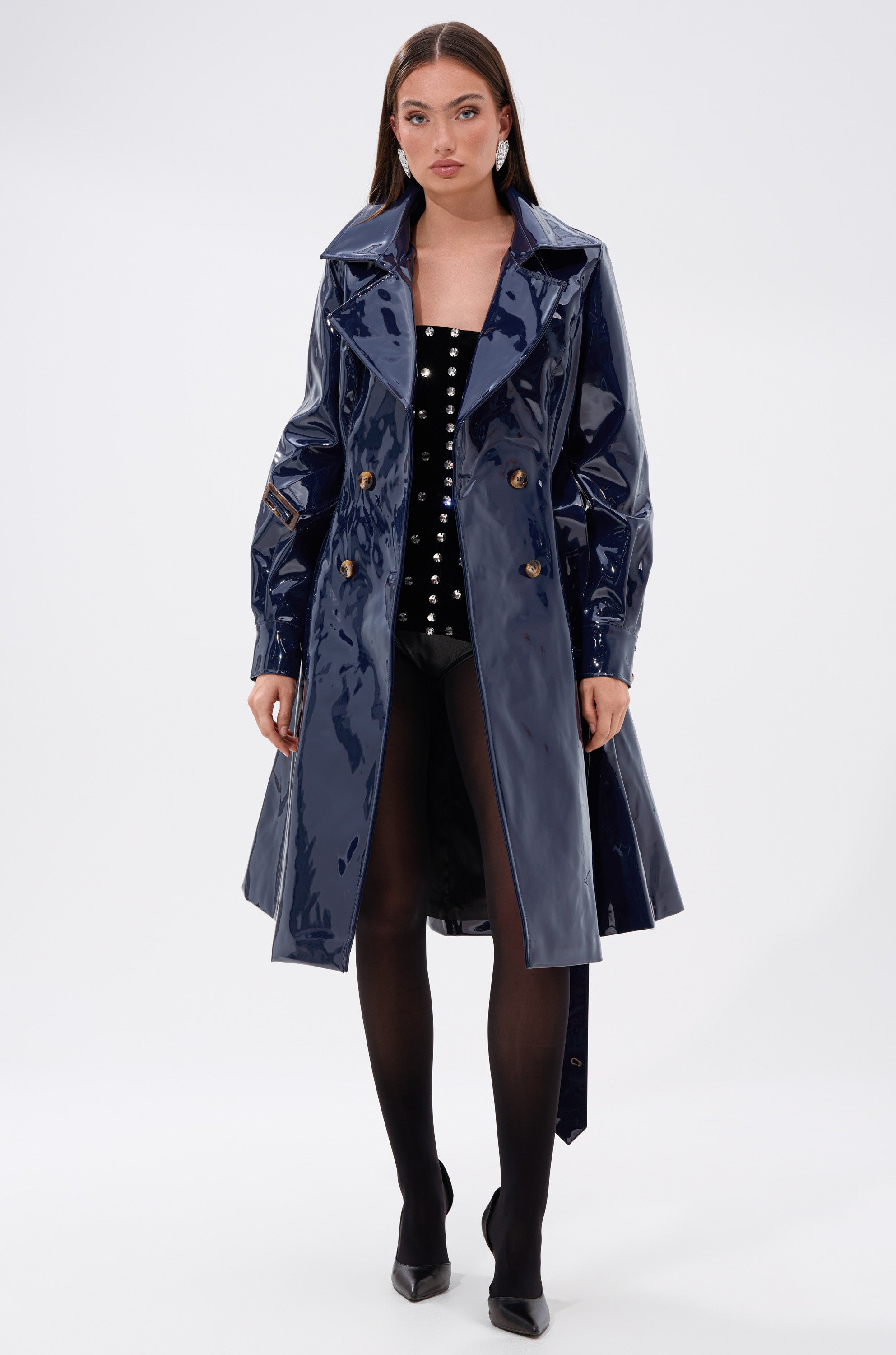 A woman faces forward in the MITICA PATENT TRENCH IN NAVY, a shiny faux patent leather coat with an adjustable waist belt, styled over a black outfit with silver studs, black tights, and heels against a plain white background.